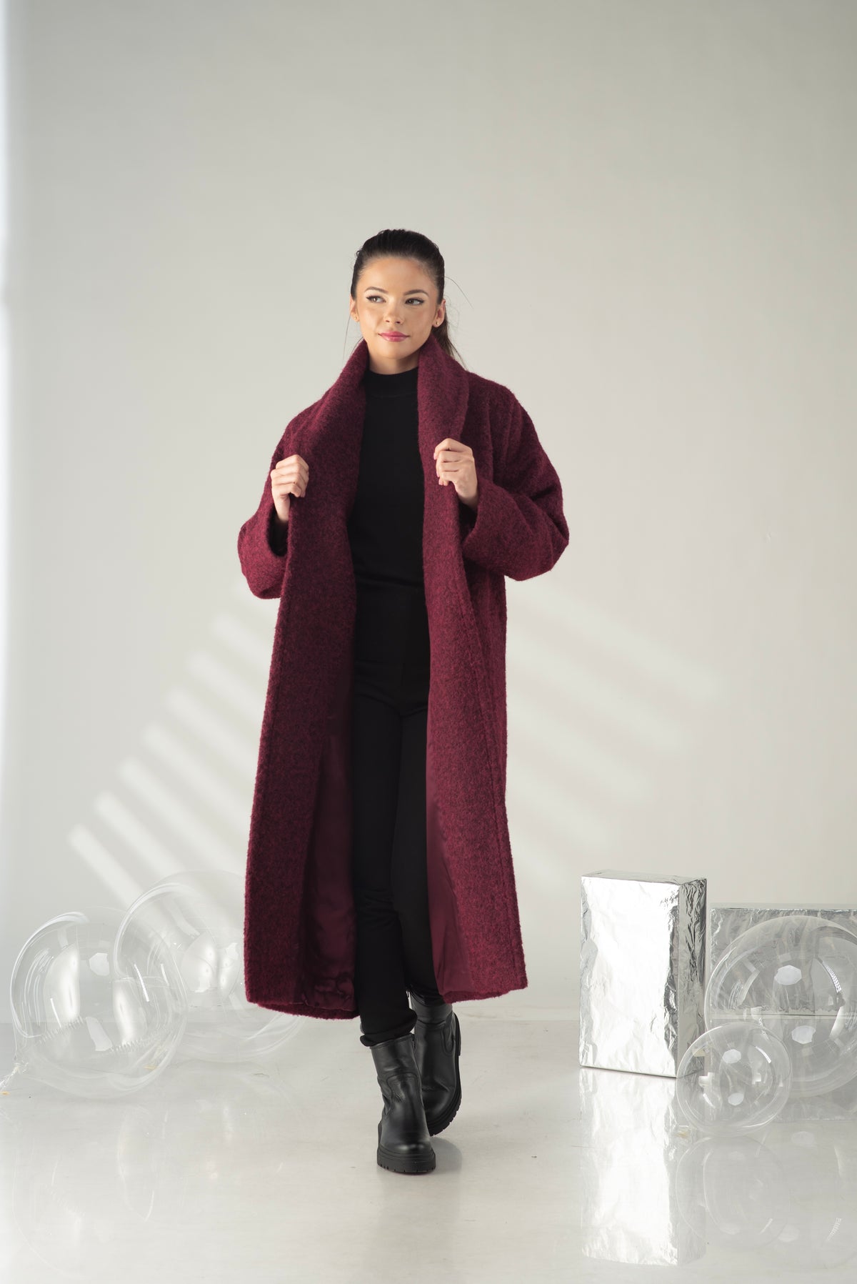 A woman modeling a long, plush maroon coat over a black outfit, standing in a bright studio with decorative clear spheres and a silver box on the floor.