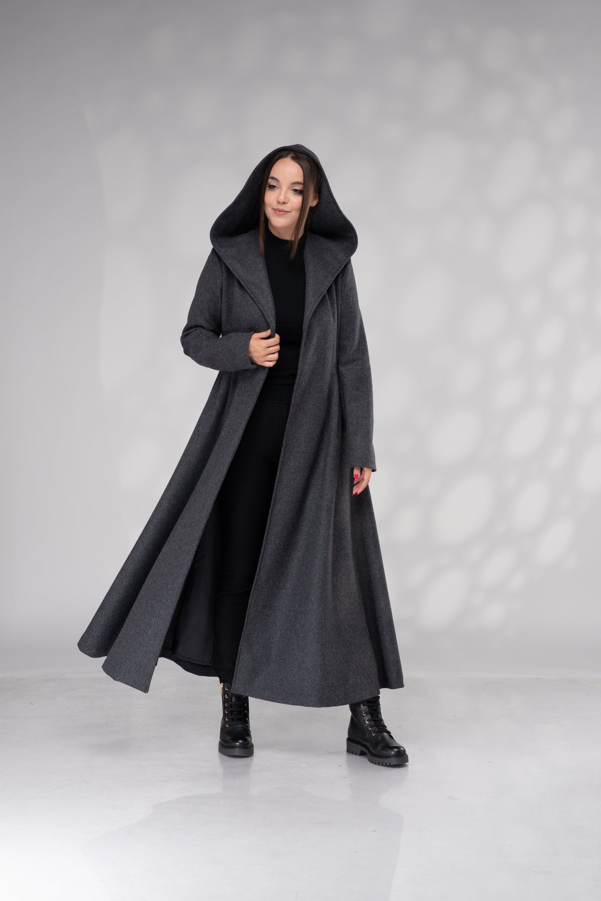 A woman modeling a long, dark gray hooded coat over a black dress and black boots, standing against a light gray studio backdrop with soft shadow patterns.