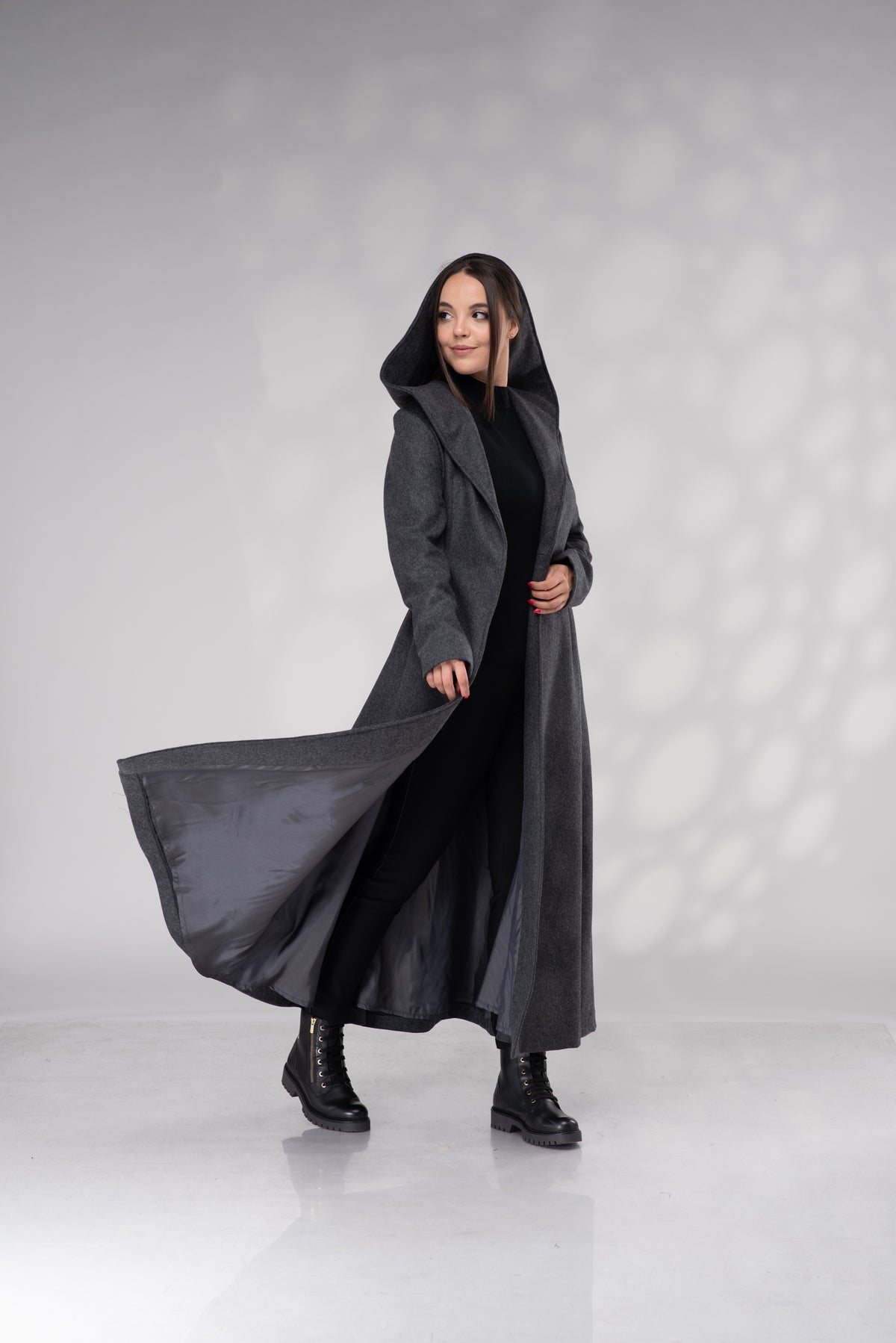 A woman modeling a long, dark gray hooded coat over a black outfit and black boots, standing in a studio with a light gray background and soft shadow patterns.