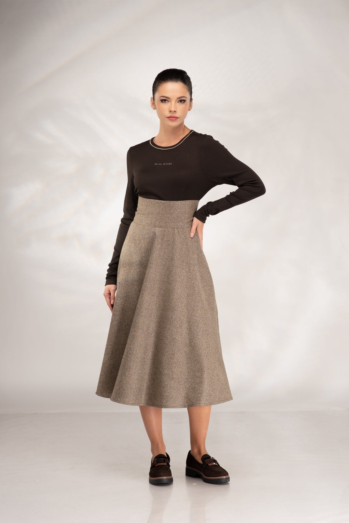 A woman modeling a two-piece outfit consisting of a dark brown long-sleeve knit top and a high-waisted, flared taupe-beige wool skirt, paired with dark brown loafers, against a light gray studio backdrop.