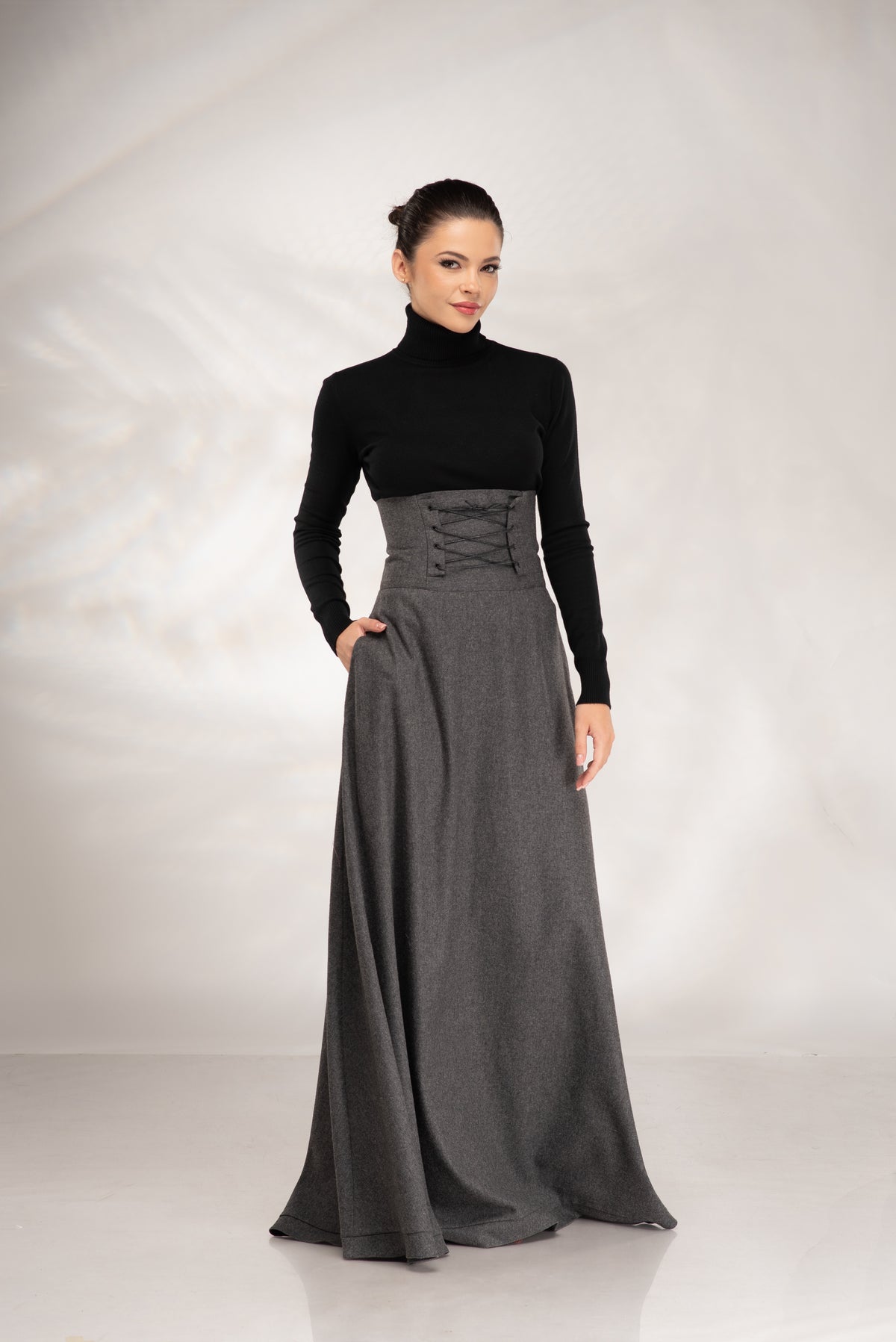 A woman modeling a long dark gray maxi dress with a black long-sleeve mock neck top, standing against a light gray studio backdrop.
