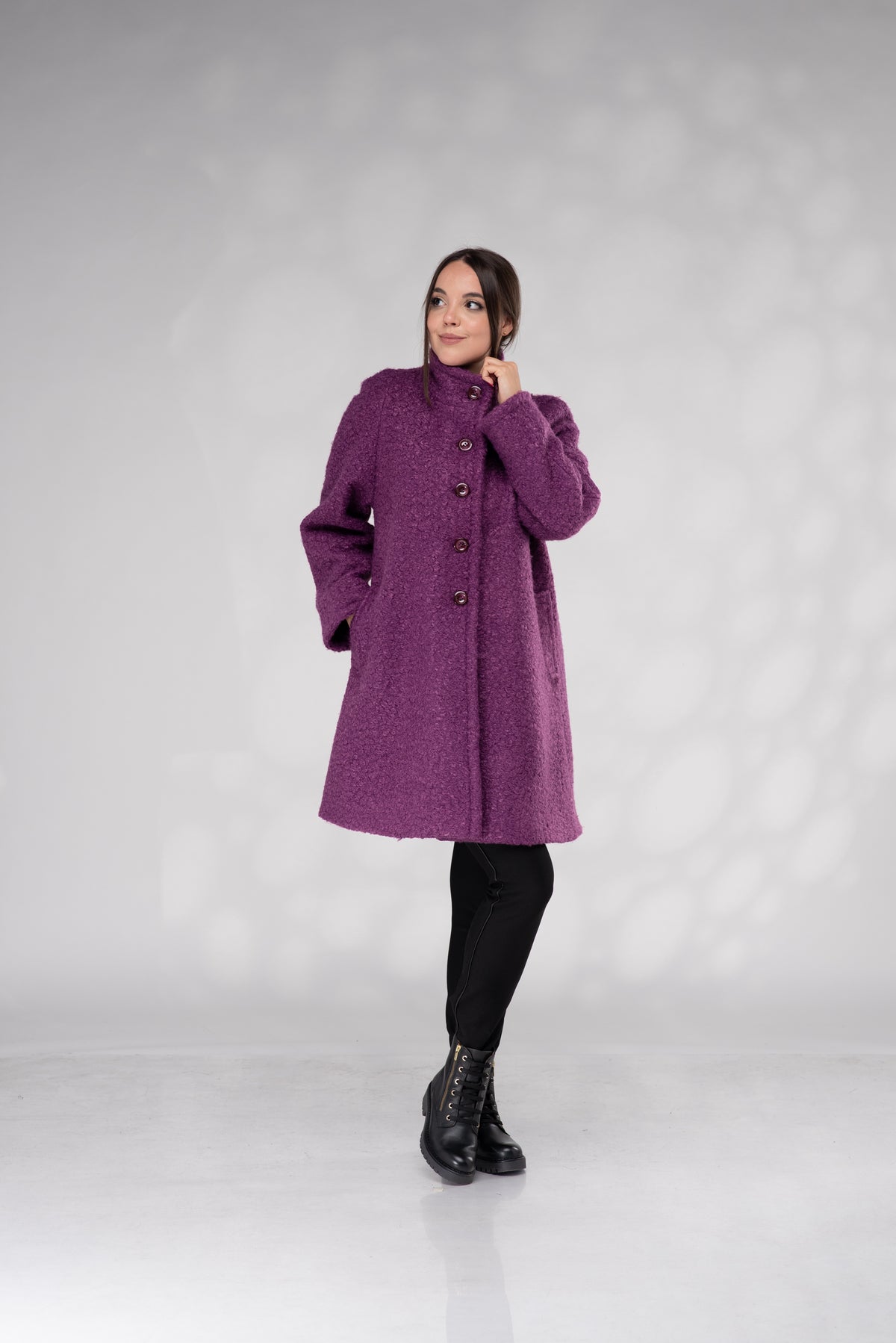 A woman modeling a textured, high-neck purple coat with dark buttons, paired with black pants and black lace-up boots, standing against a light gray studio backdrop.