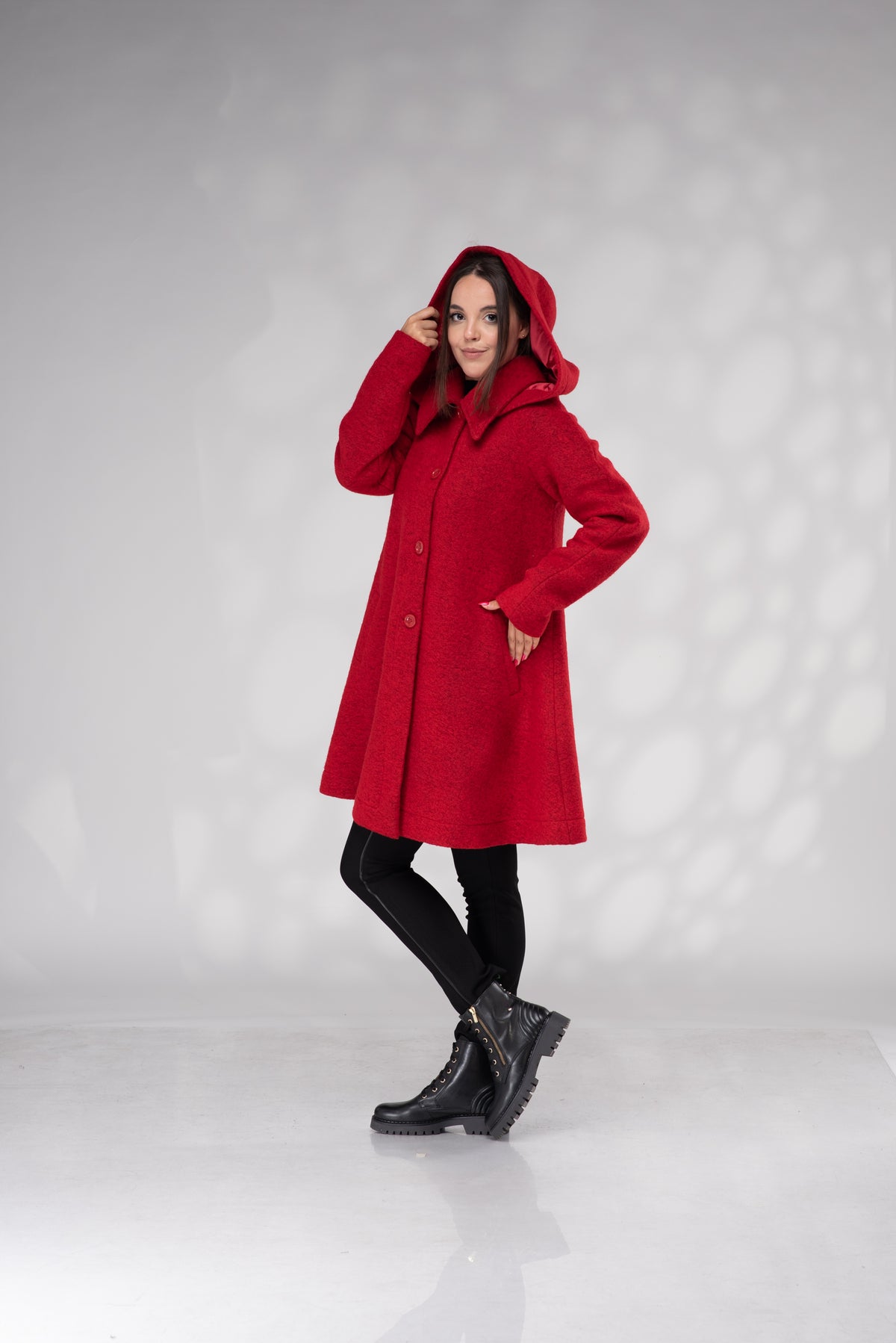 A woman modeling a bright red hooded coat with black leggings and black boots, standing against a light gray studio backdrop with soft circular shadows.