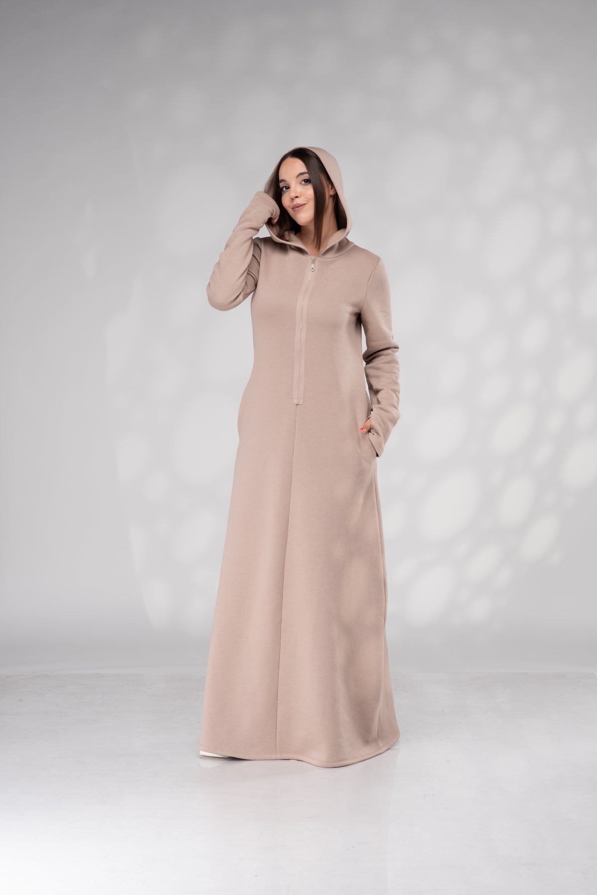 A woman modeling a long, floor-length hooded abaya in a dusty rose-beige color, standing against a light gray studio backdrop with soft shadow patterns.
