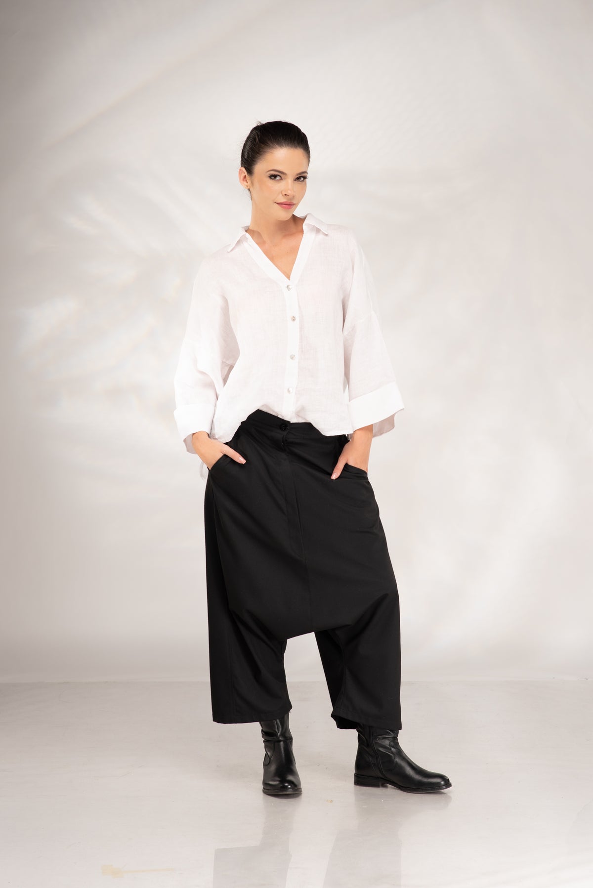 A woman modeling a white long-sleeve button-up shirt and black wide-leg harem pants with black ankle boots, standing against a light gray studio backdrop.