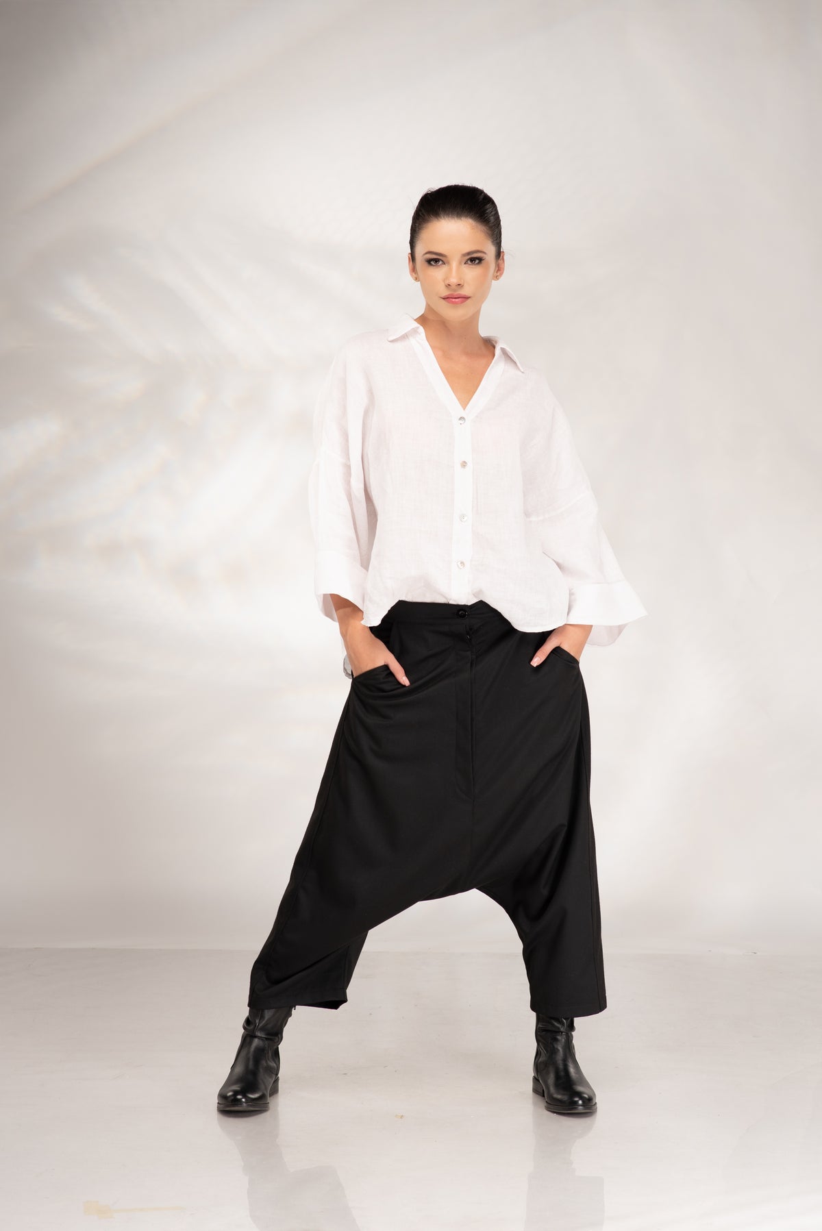 A model wearing a white long-sleeve button-up shirt and black baggy harem-style pants, standing against a white studio backdrop.