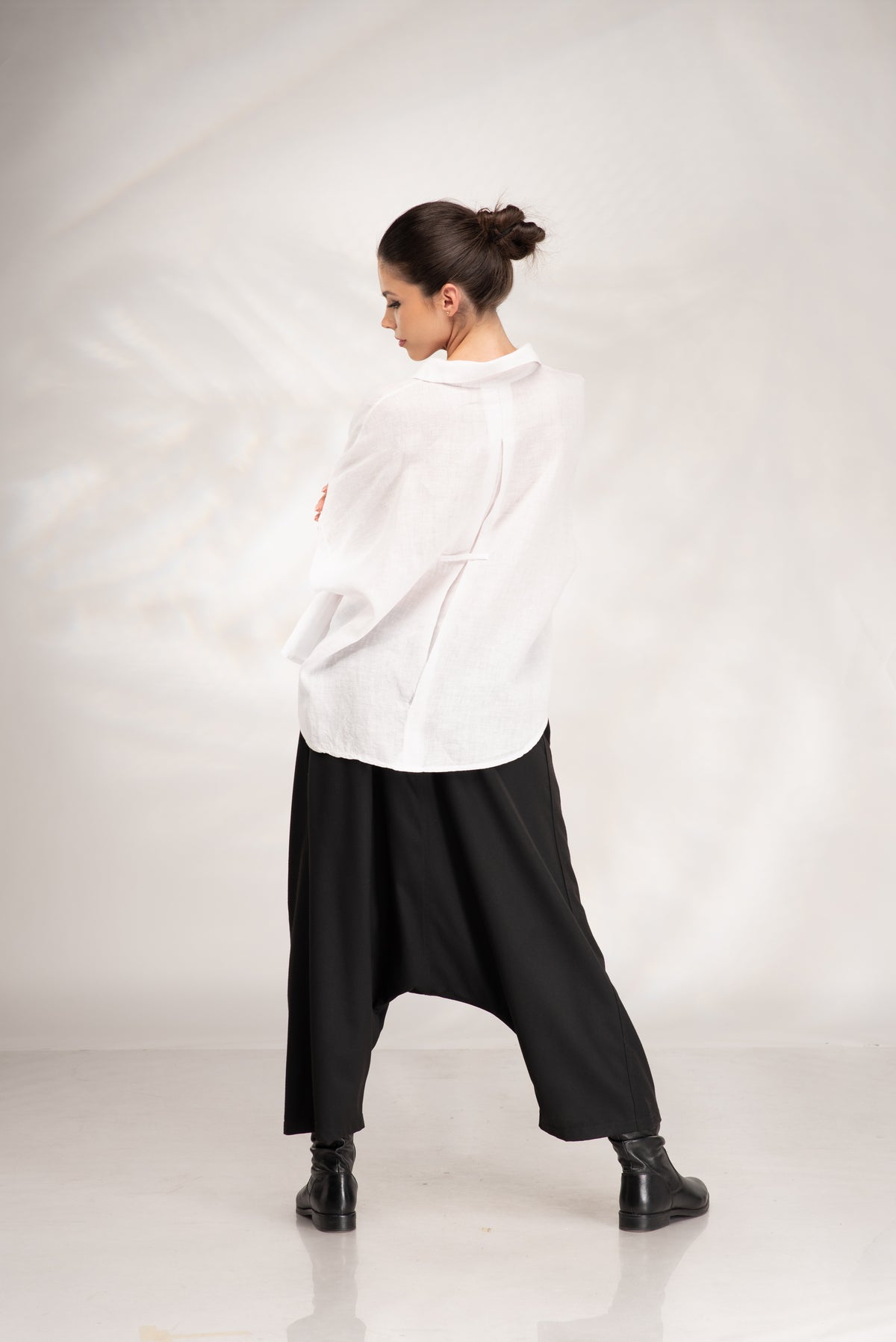 A woman wearing a loose white long-sleeve shirt and black harem-style pants with black boots, photographed from the back against a light gray studio backdrop.