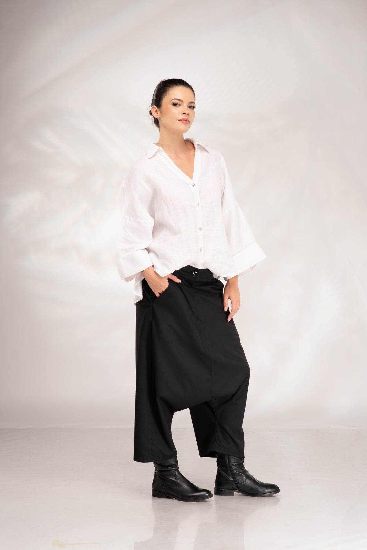 A woman modeling a white long-sleeve button-up shirt and black baggy wide-leg pants with black leather boots, standing against a light gray studio backdrop.