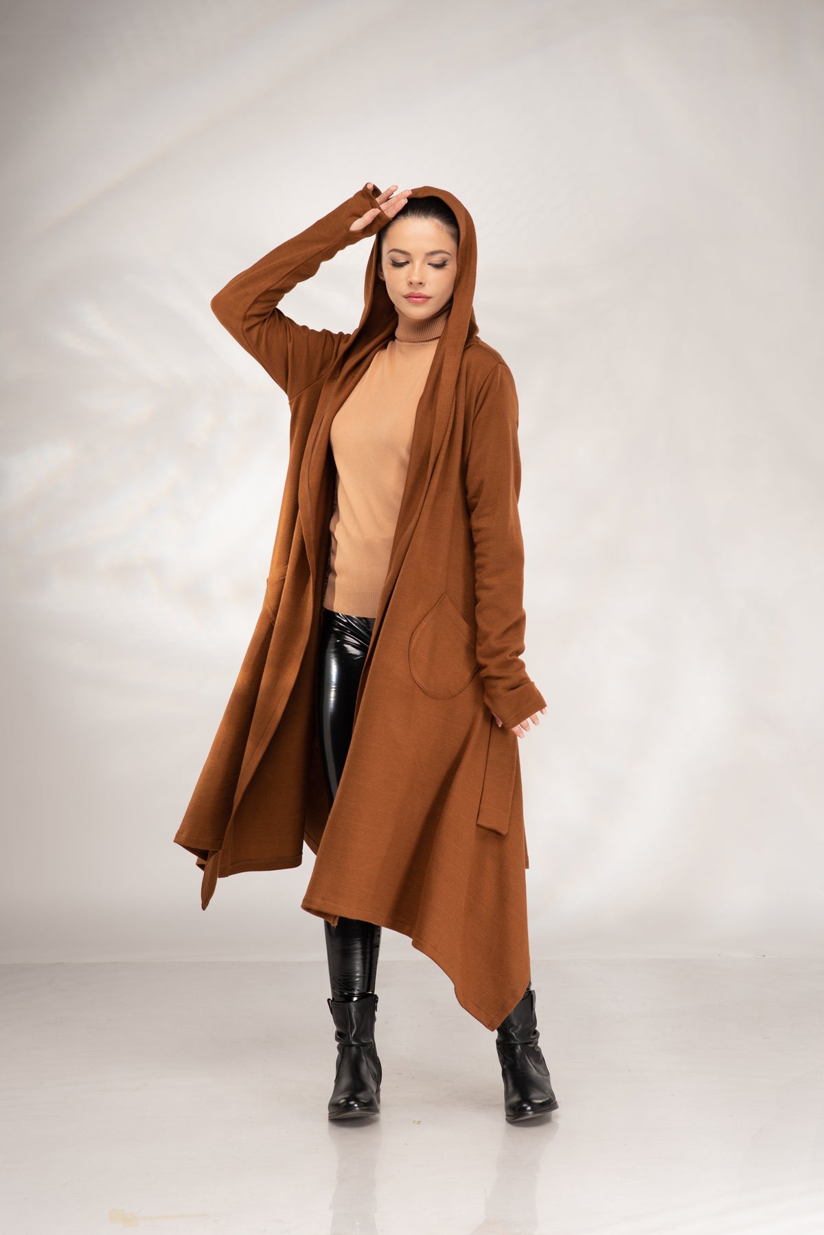 A woman modeling a long, open-front brown hooded cardigan over a beige top and black pants, standing against a light gray studio backdrop.