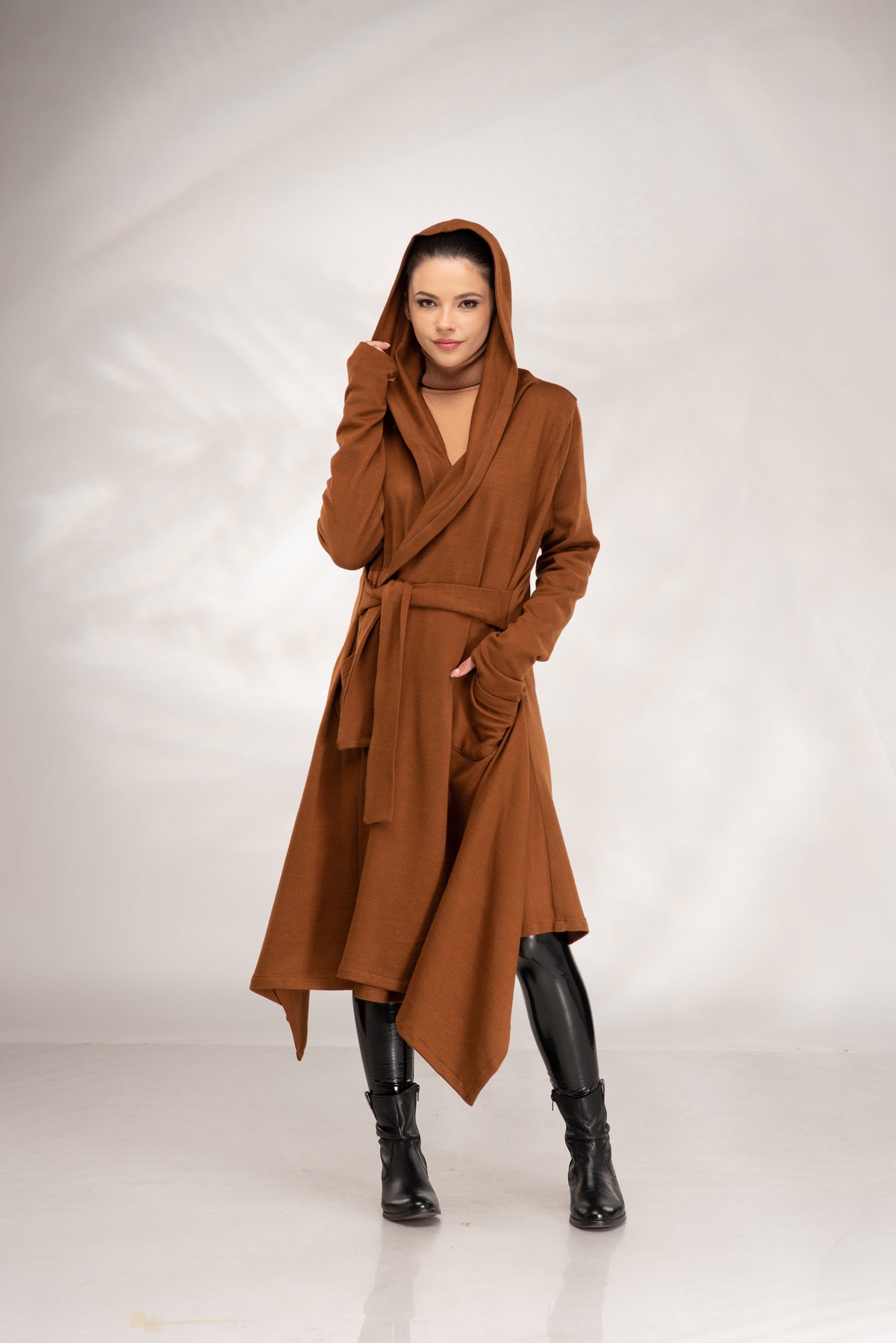 A woman modeling a long brown hooded wrap coat with a tie belt, worn over a beige top and black knee-high boots, against a light gray studio backdrop.