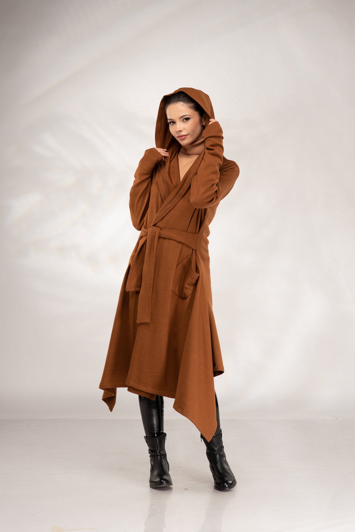 A woman modeling a brown hooded wrap coat with a matching belt, paired with black ankle boots, against a light gray studio backdrop.