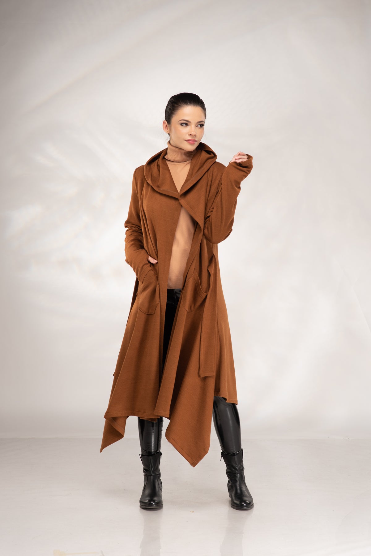 A woman modeling a long, asymmetrical brown hooded coat over a beige top and black pants and boots, standing against a light gray studio backdrop.