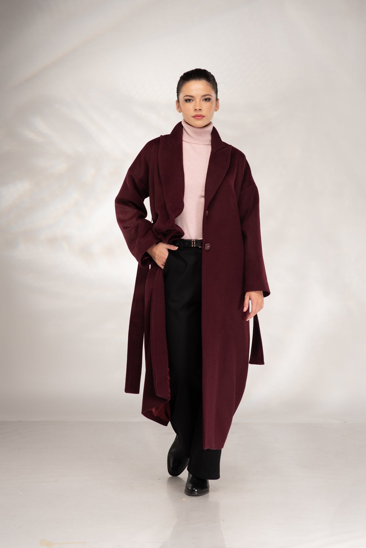 A woman modeling a long, open-front burgundy coat over a light pink turtleneck and black trousers, standing against a light gray studio backdrop.
