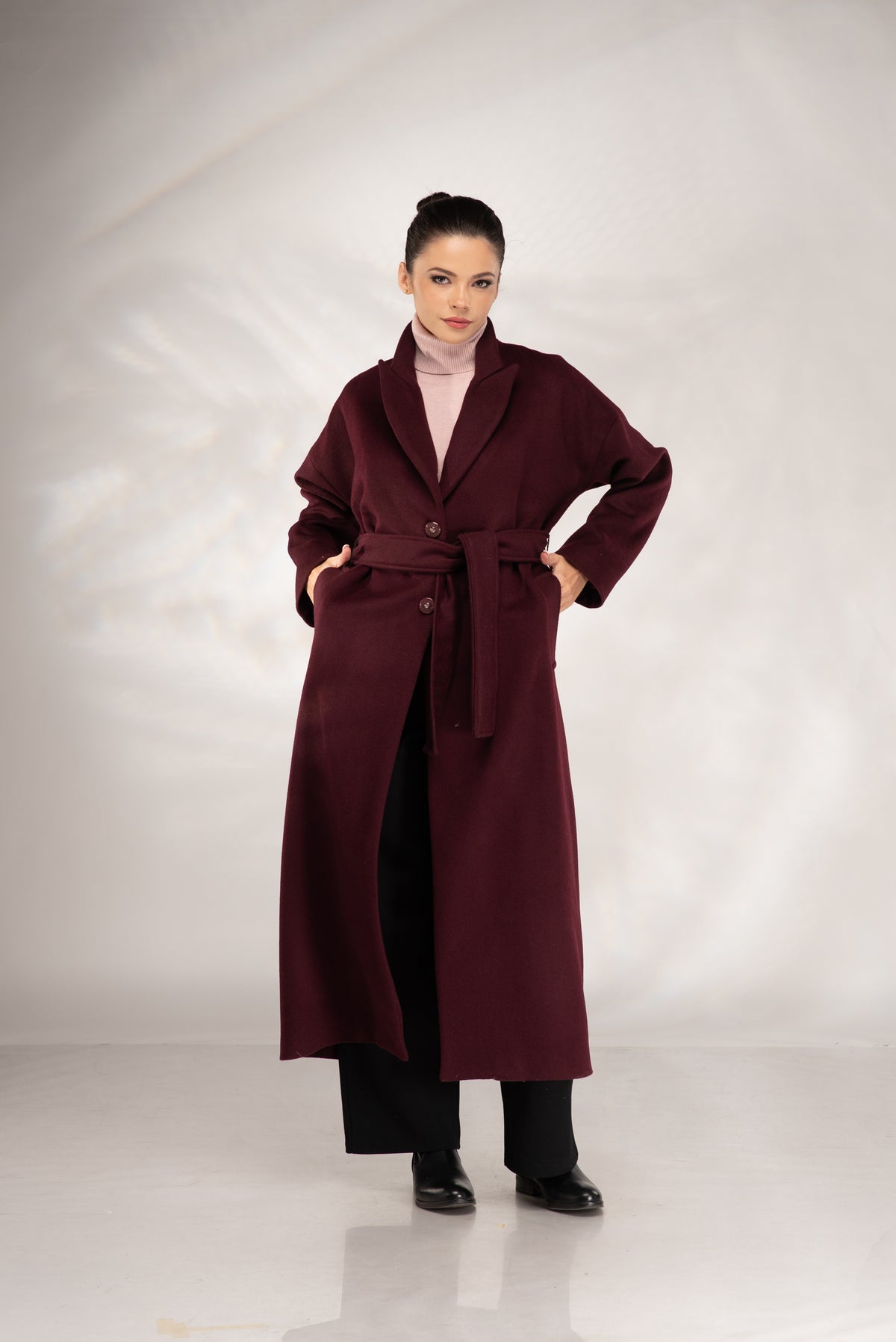 A woman modeling a long burgundy belted coat over a light pink turtleneck and black pants, standing against a light gray studio backdrop.