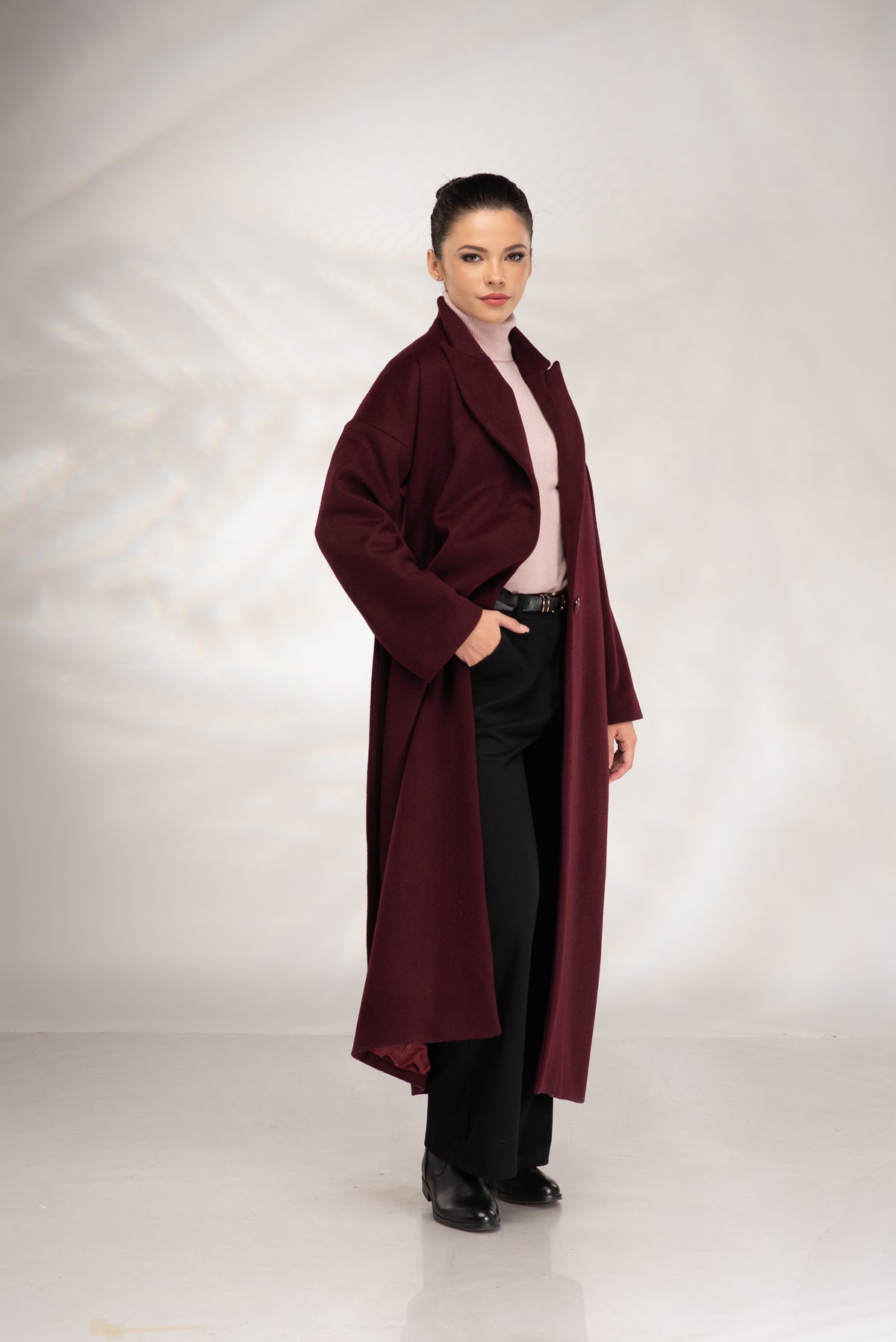 A woman modeling a long burgundy wool-blend coat over a light pink top and black trousers, standing against a white studio backdrop.
