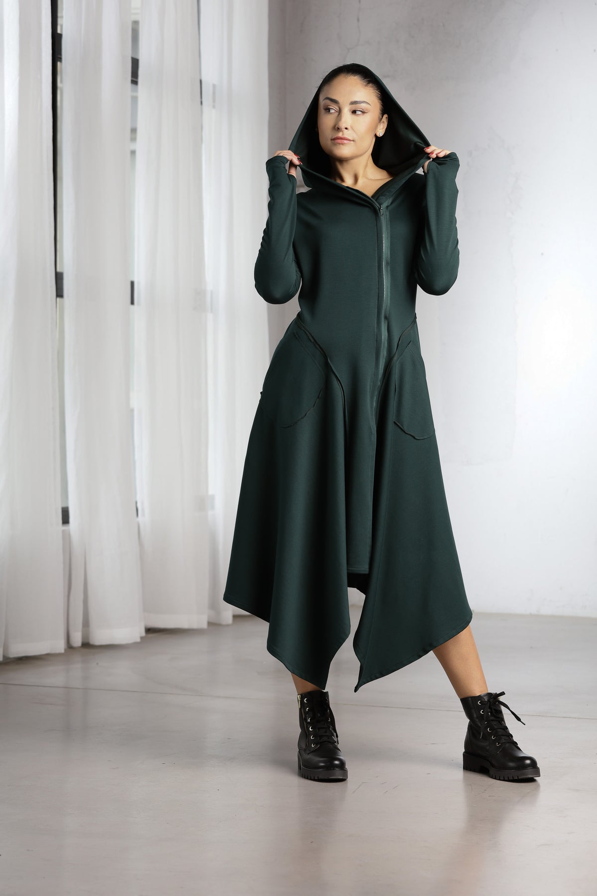 A woman models a dark forest green asymmetrical zip-up hoodie dress with a high-low hem, styled with black combat boots in a bright, modern studio setting.