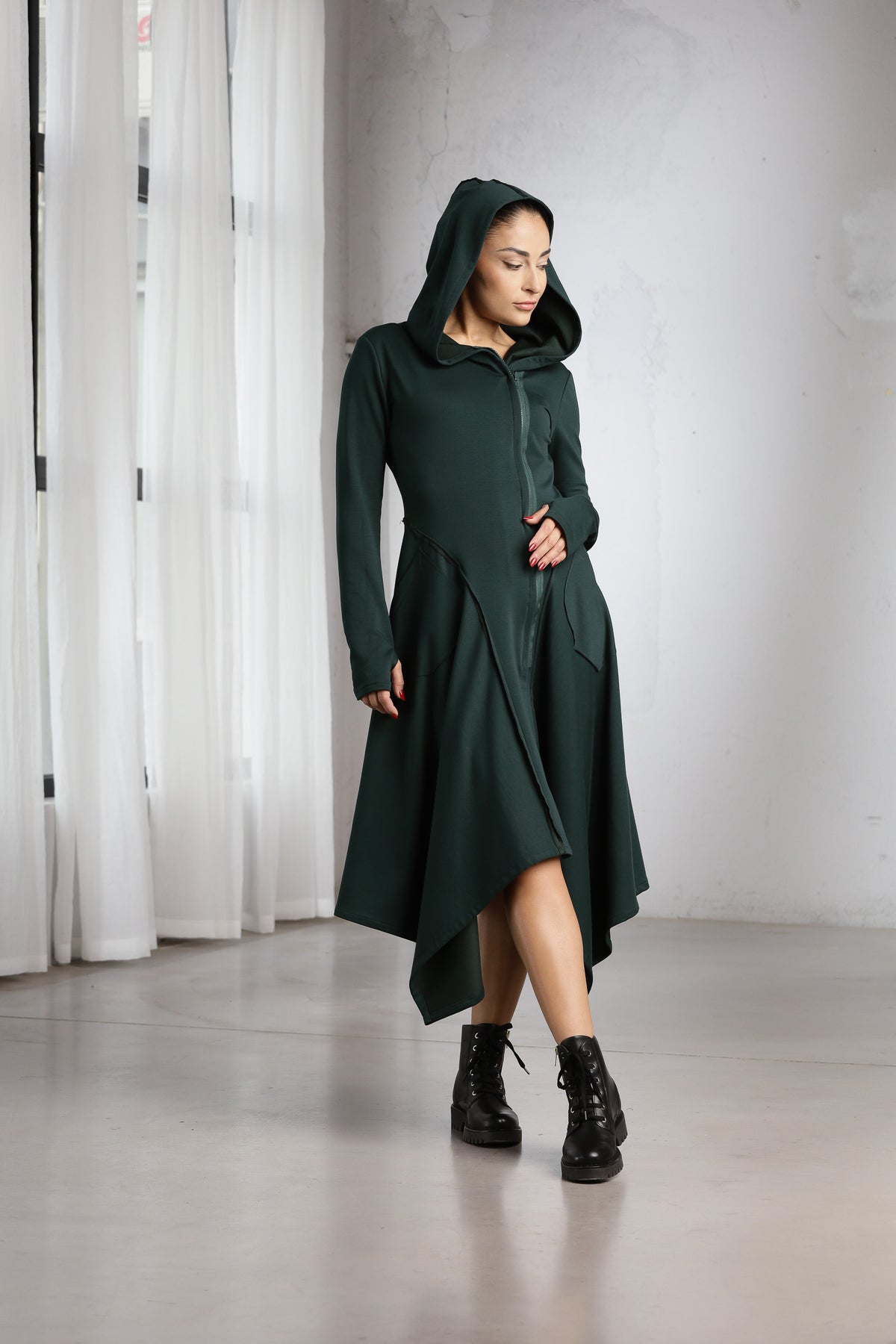 A woman wearing a dark forest green hooded long-sleeve dress with an asymmetrical draped hem and black lace-up ankle boots, standing in a minimalist studio with white curtains and a light gray floor.