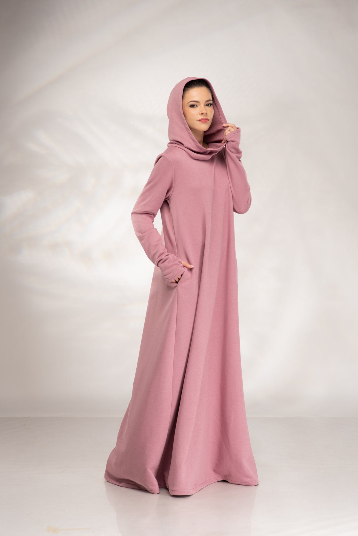 A woman modeling a long, flowing dusty pink abaya with a matching headscarf, standing against a light gray studio backdrop.