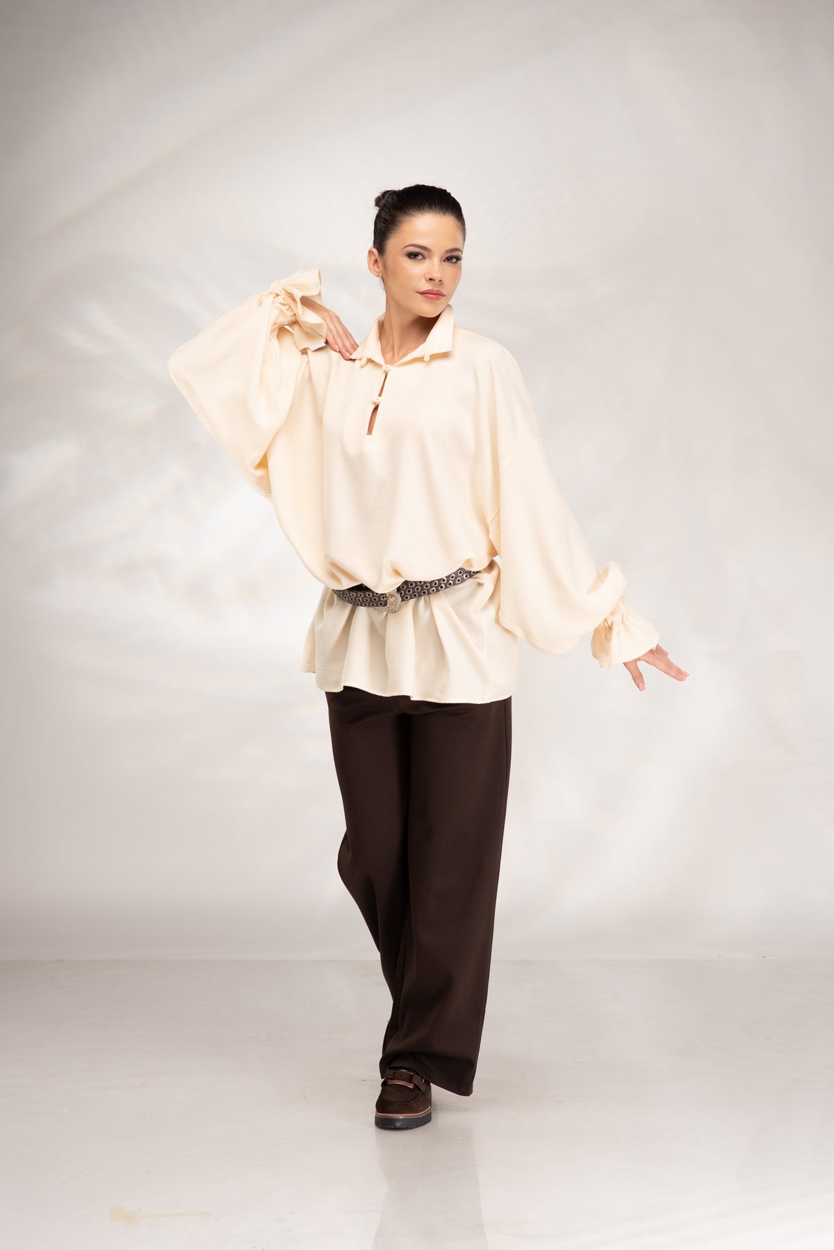 A woman modeling a cream-colored long-sleeve blouse with a half-zip neckline and a dark brown sash, paired with dark brown wide-leg pants, against a light gray studio backdrop.