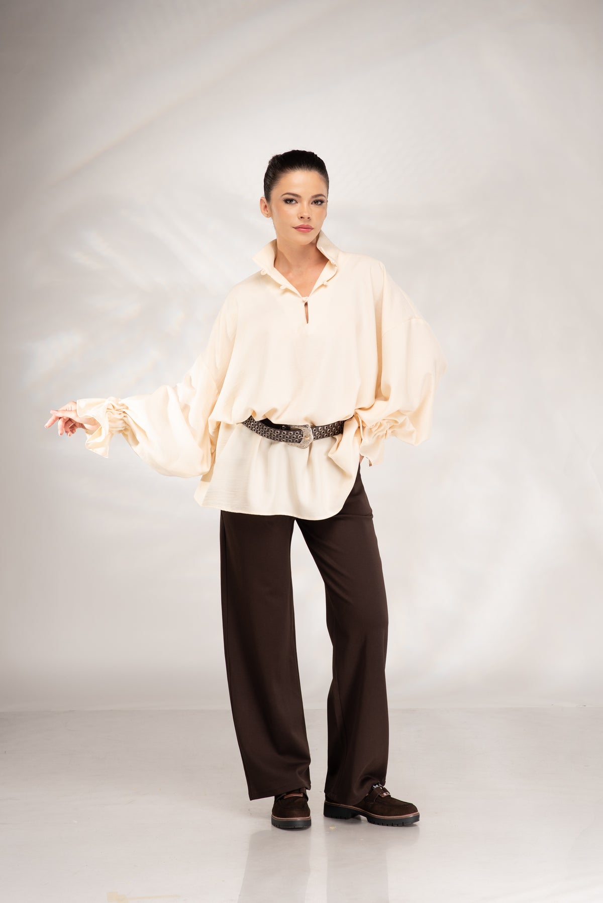 A model stands against a light gray studio backdrop wearing a cream-colored, oversized long-sleeve blouse with dramatic ruffled sleeves, dark brown wide-leg trousers, and dark brown shoes.