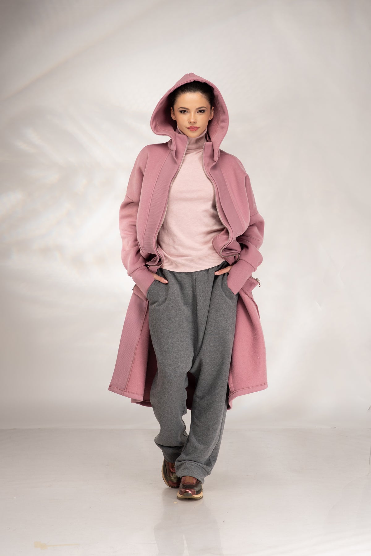 A model wearing a dusty pink hooded coat over a light pink top, gray sweatpants, and brown shoes, standing against a light gray studio backdrop.