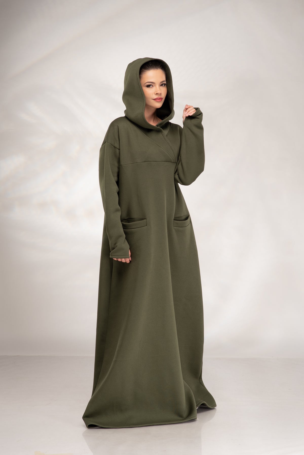 A woman modeling a long, loose-fitting olive green hooded dress with large front patch pockets, photographed against a light gray studio backdrop.