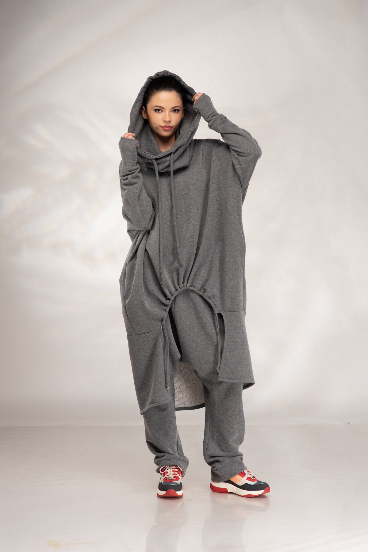 A model wearing a loose-fitting, oversized gray hooded sweatshirt and matching sweatpants, paired with red, white, and black sneakers, standing against a light gray studio backdrop.