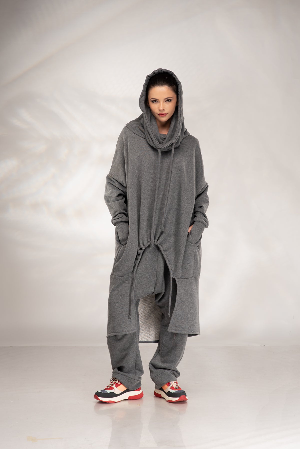 A model wearing a long, oversized gray hooded sweatshirt with a drawstring waist, matching gray sweatpants, and red, black, and white sneakers, standing against a light gray studio backdrop.