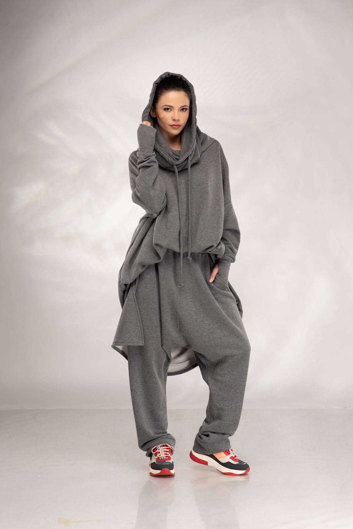 A model wearing a loose-fitting, oversized gray hooded sweatshirt and matching sweatpants, styled with colorful sneakers, standing against a light gray studio backdrop.