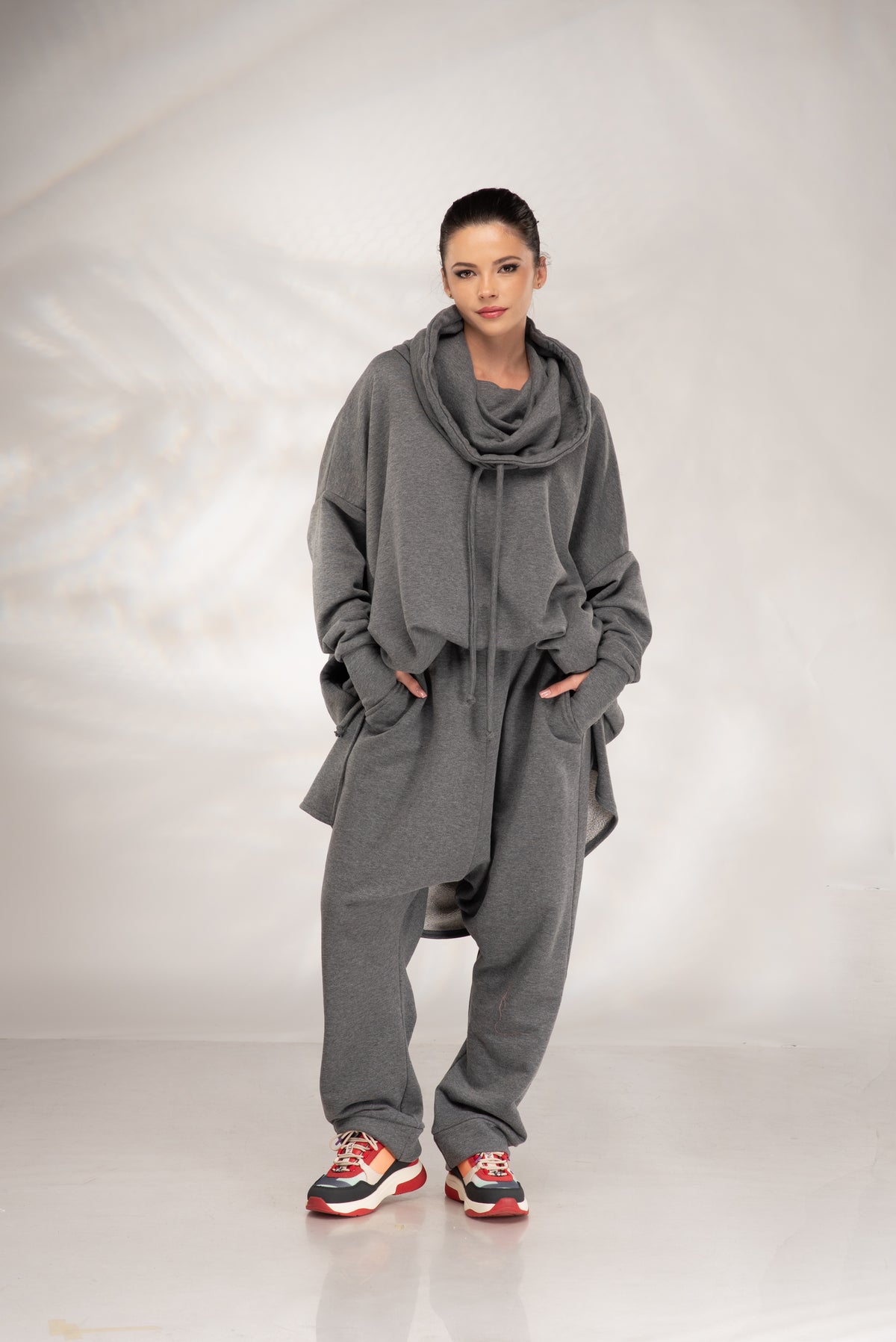A model wearing a matching gray lounge set consisting of a draped hoodie and sweatpants, styled with colorful sneakers, against a light gray studio backdrop.