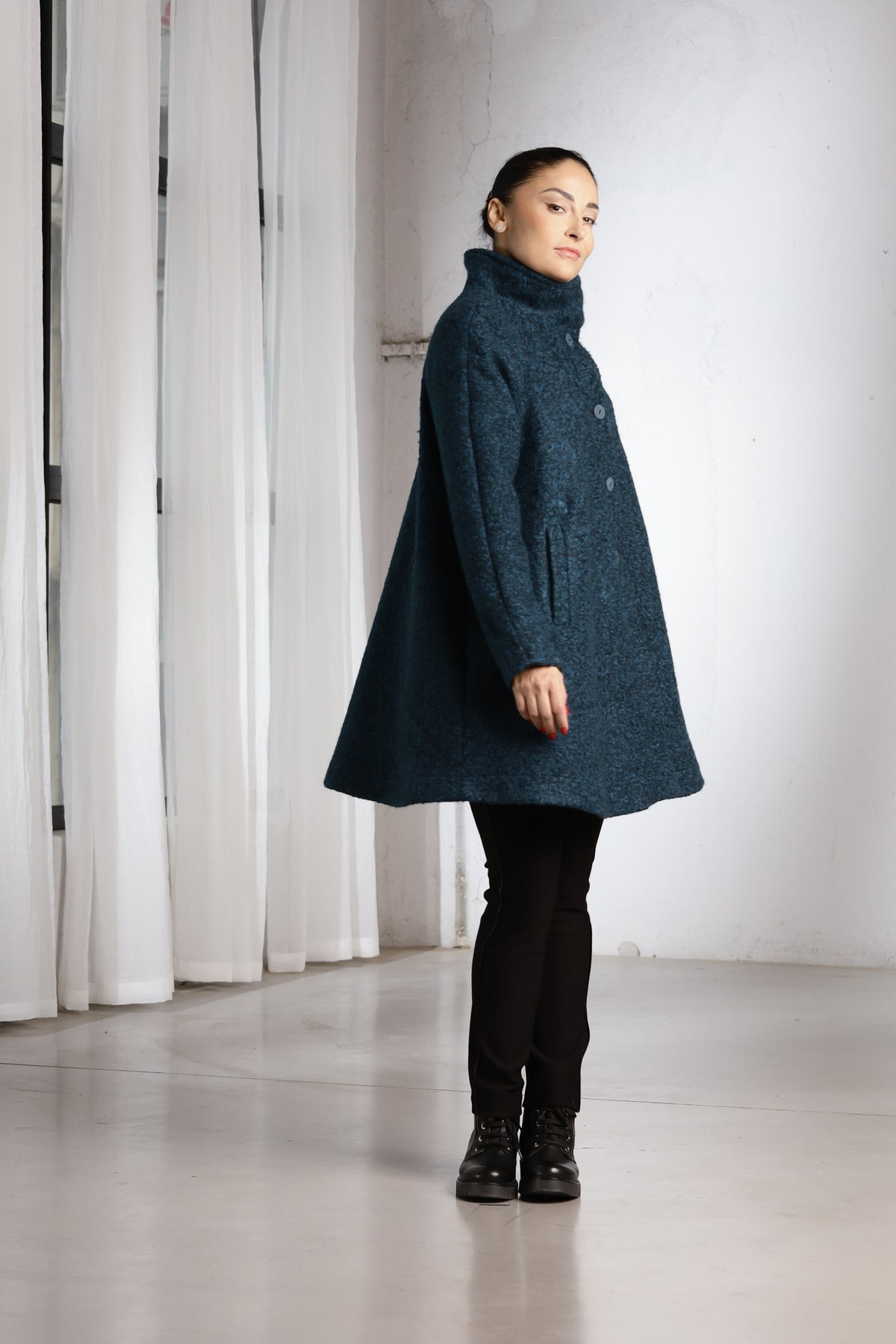 A woman modeling a dark teal textured wool coat with a high mock neck, paired with black pants and boots, standing in a minimalist studio with white curtains.