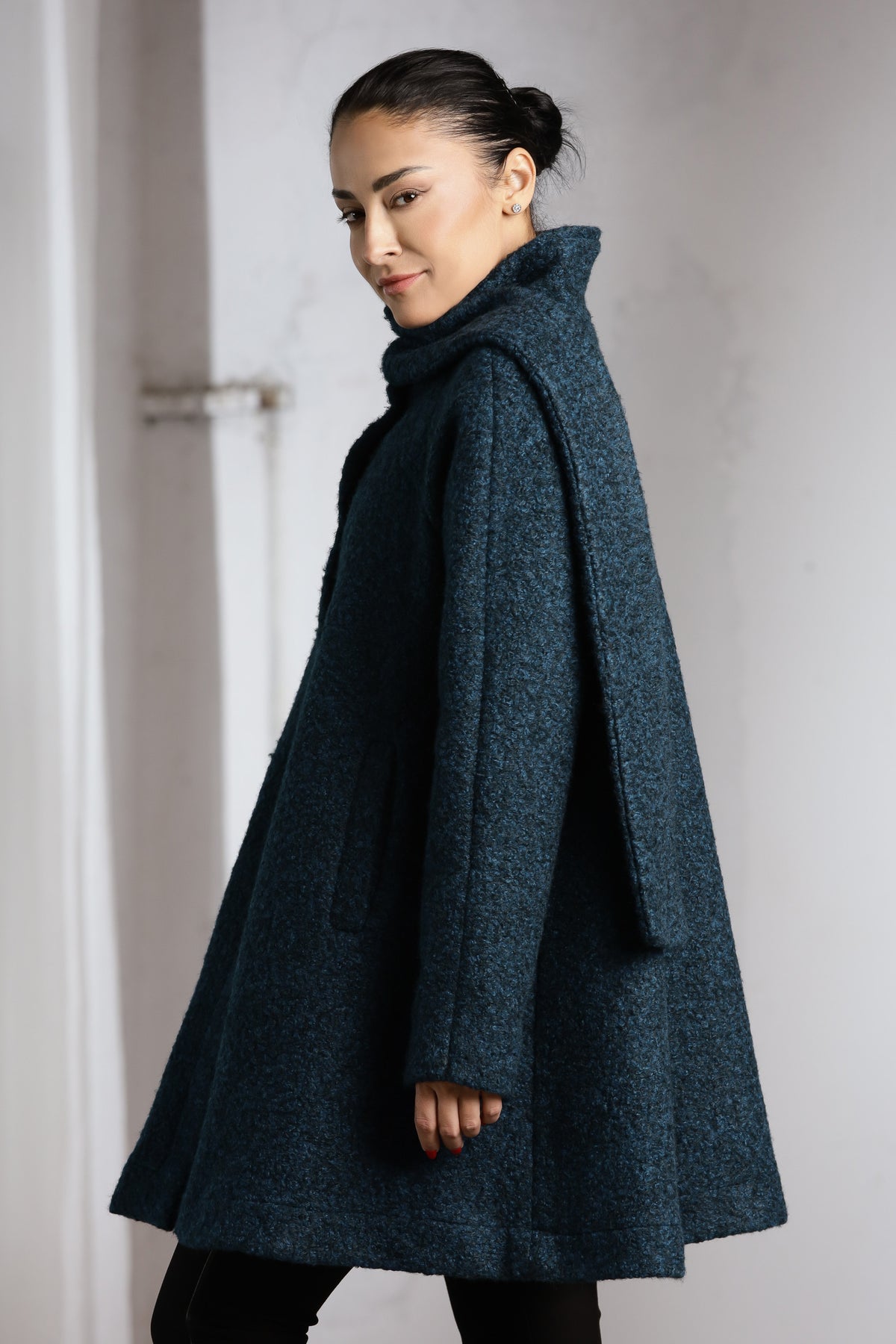 A woman wearing a dark teal textured coat with a high collar, shown from a side-back angle against a light gray wall.