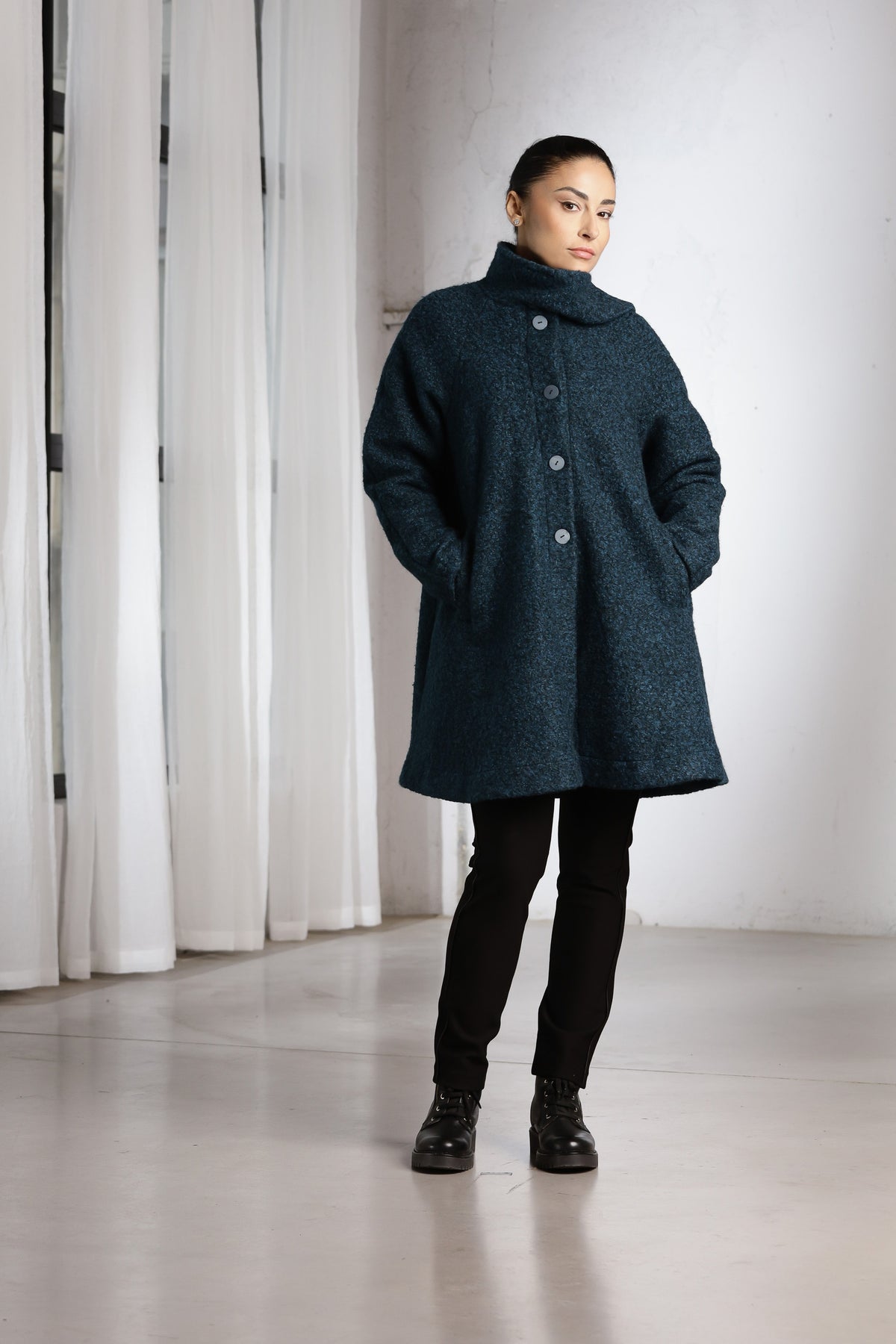 A woman modeling a dark teal textured coat with a high collar and front buttons, paired with black pants and boots, standing in a minimalist room with white curtains.