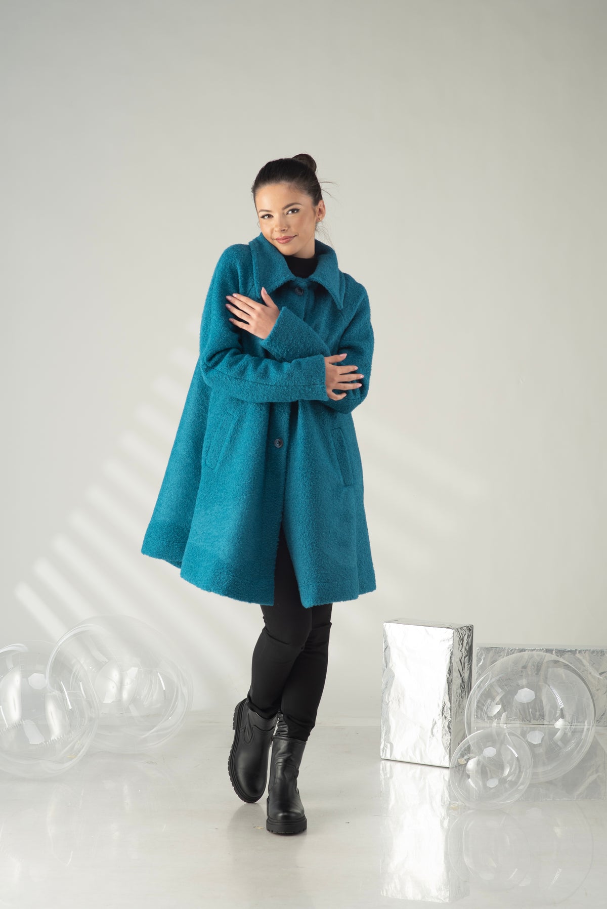 A woman wearing a teal blue wool coat over a black turtleneck and black boots, posing in a studio setting with decorative clear plastic spheres and a silver box.