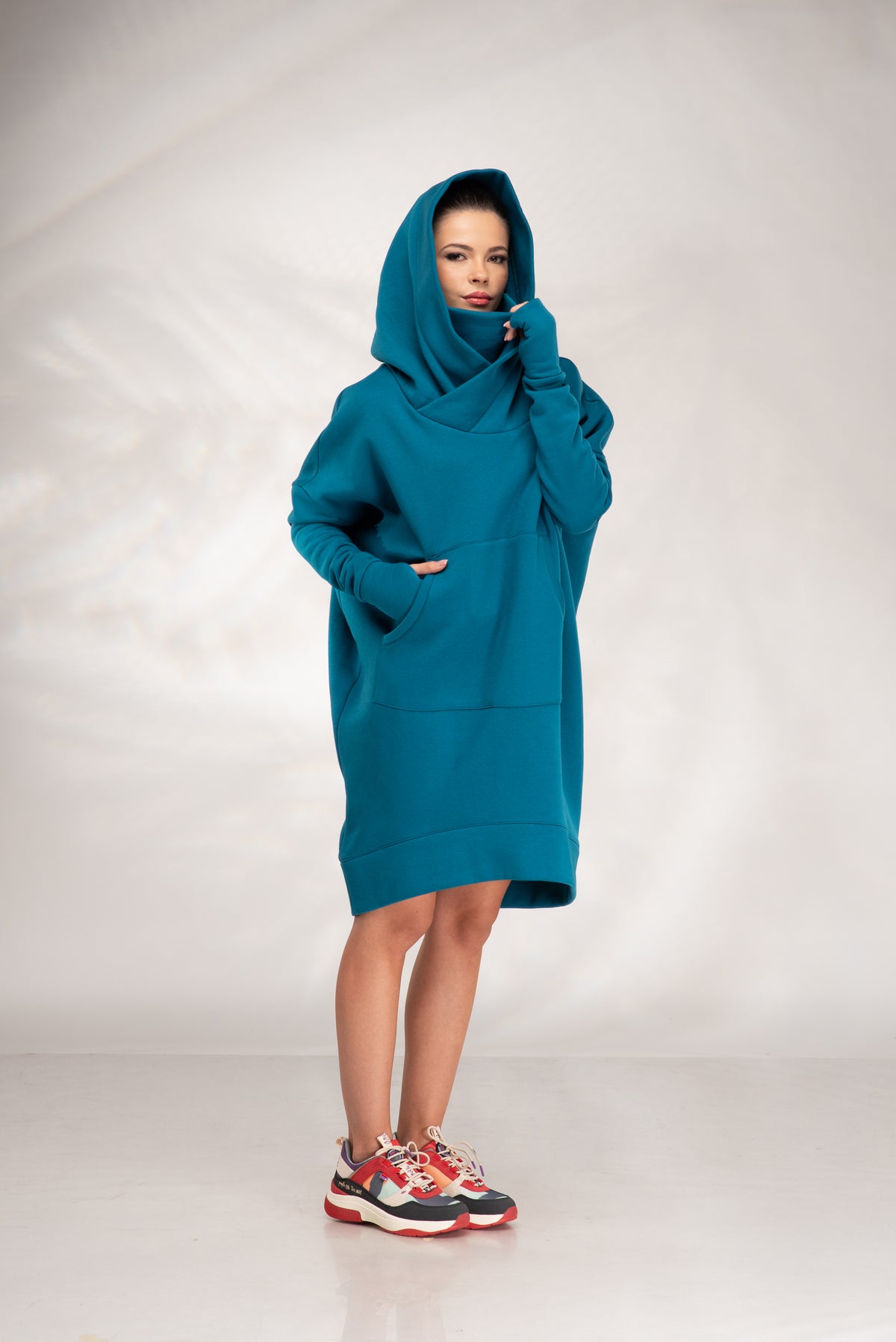 A model wearing a teal oversized hooded sweatshirt dress with a high folded collar, standing against a light gray studio backdrop.