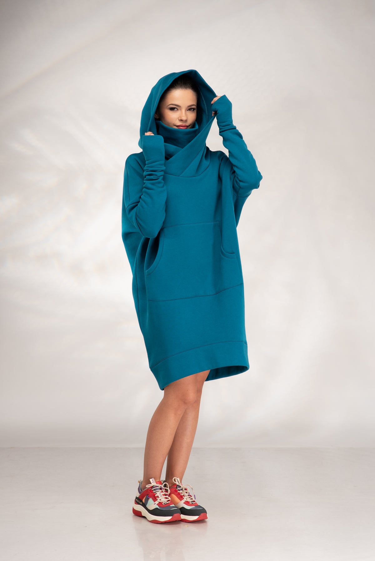 A woman modeling an oversized teal blue-green hooded sweatshirt dress with a high folded collar, front pockets, and a relaxed silhouette, styled with red and black sneakers against a light gray studio backdrop.