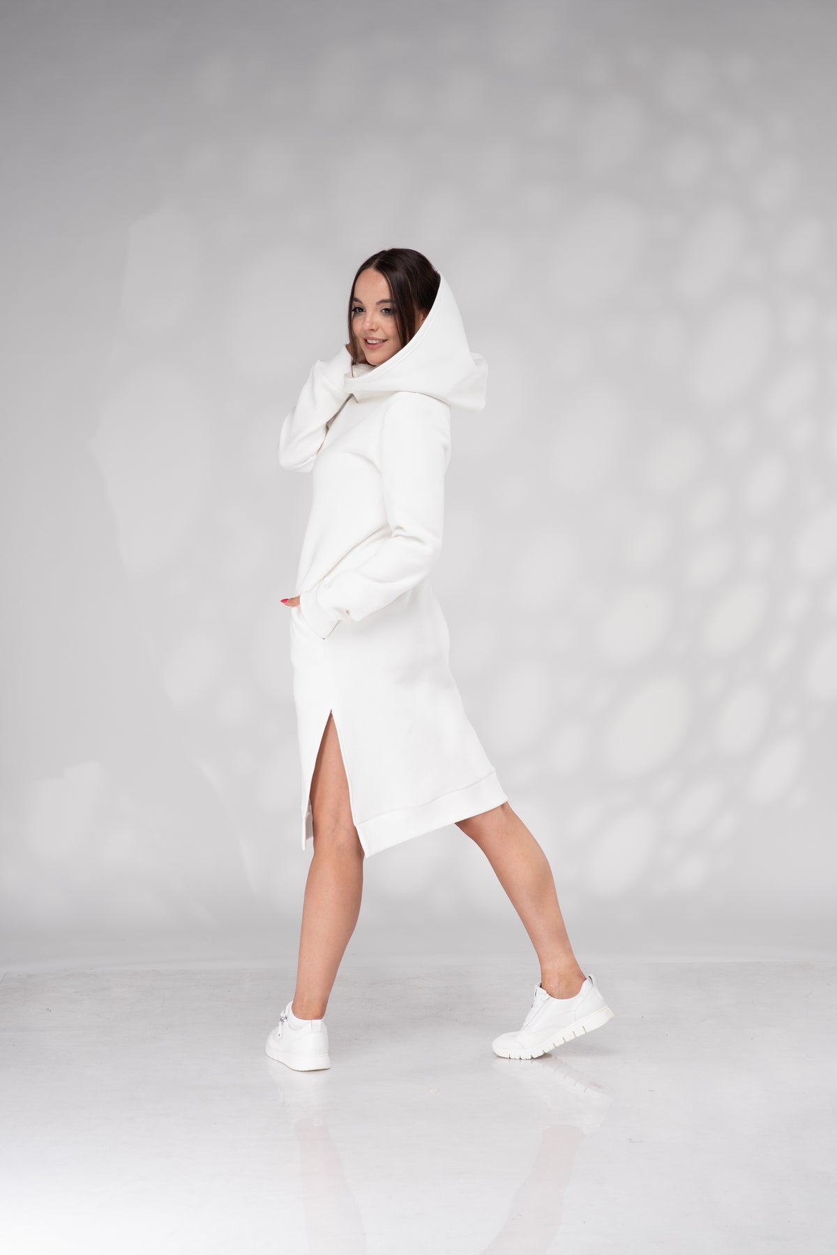 A woman modeling a white hooded longline coat or dress with a thigh-high slit, paired with white sneakers, against a light gray studio backdrop with soft circular light patterns.