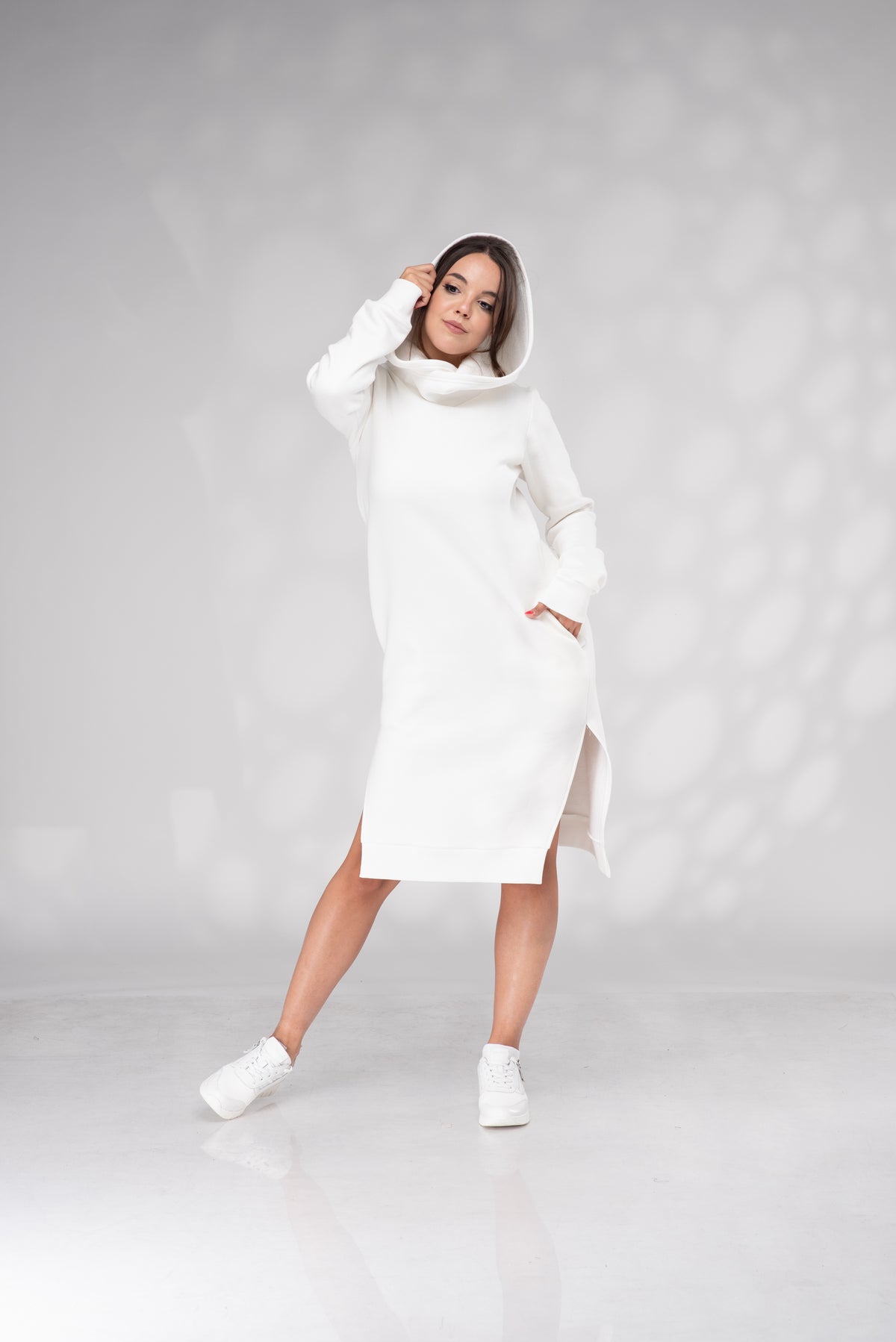 A woman modeling a white hooded knit sweater dress with a high cowl neck and side slits, styled with white sneakers against a light gray studio backdrop with soft circular light patterns.