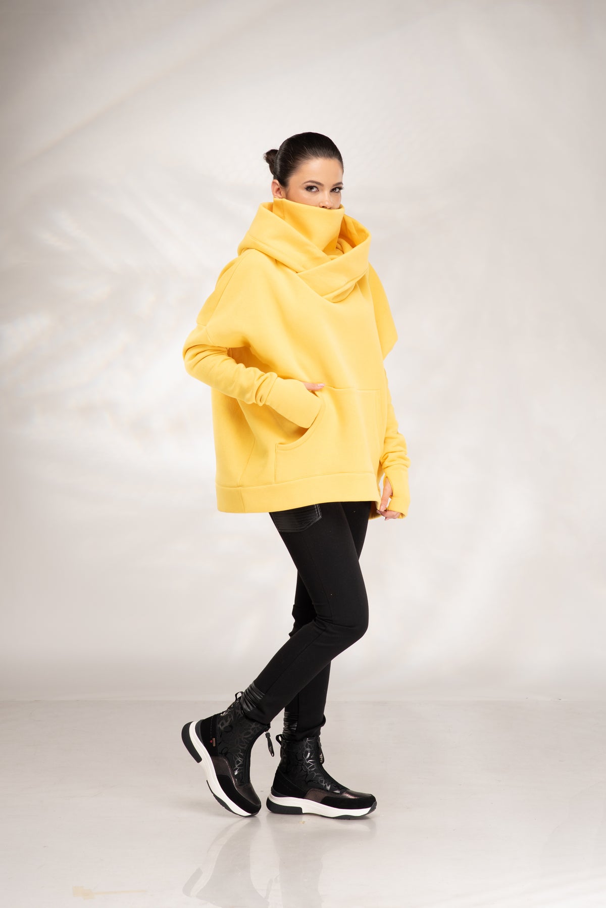 A woman modeling an oversized bright yellow hooded sweatshirt with a high folded collar, paired with black pants and black boots, against a light gray studio backdrop.
