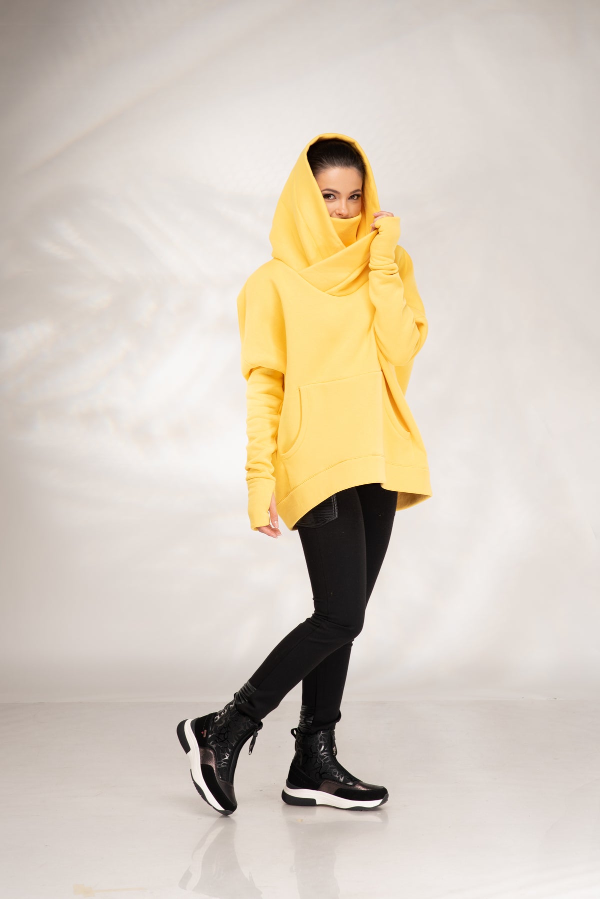 A woman modeling a bright yellow oversized cowl-neck hoodie with black skinny pants and black high-top sneakers, standing against a light gray studio backdrop.
