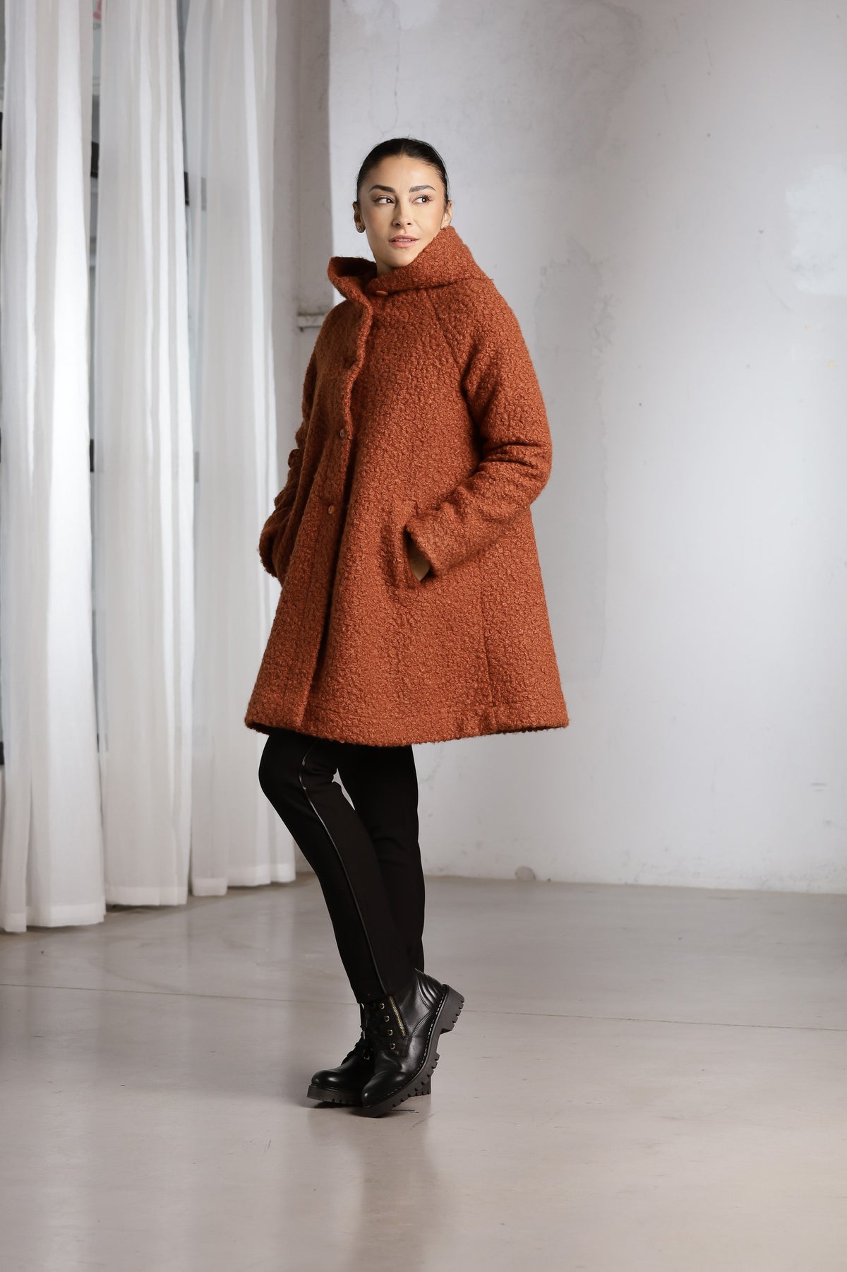 A woman modeling a textured, high-neck brown teddy coat, paired with black pants and boots, standing in a minimalist studio with white curtains and a white wall.
