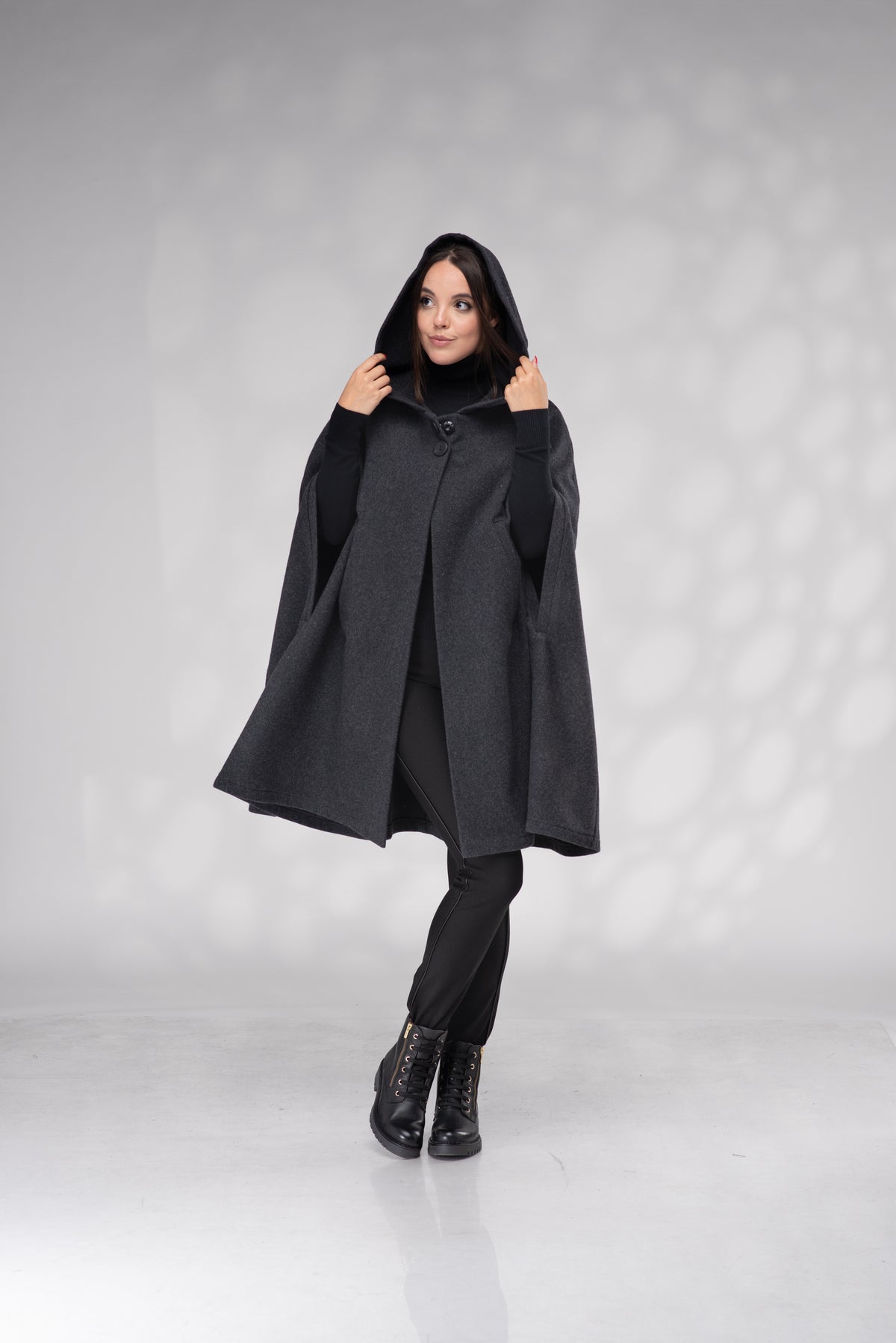 A woman modeling a dark gray hooded cape coat over a black outfit, standing against a light gray studio backdrop with soft shadow patterns.