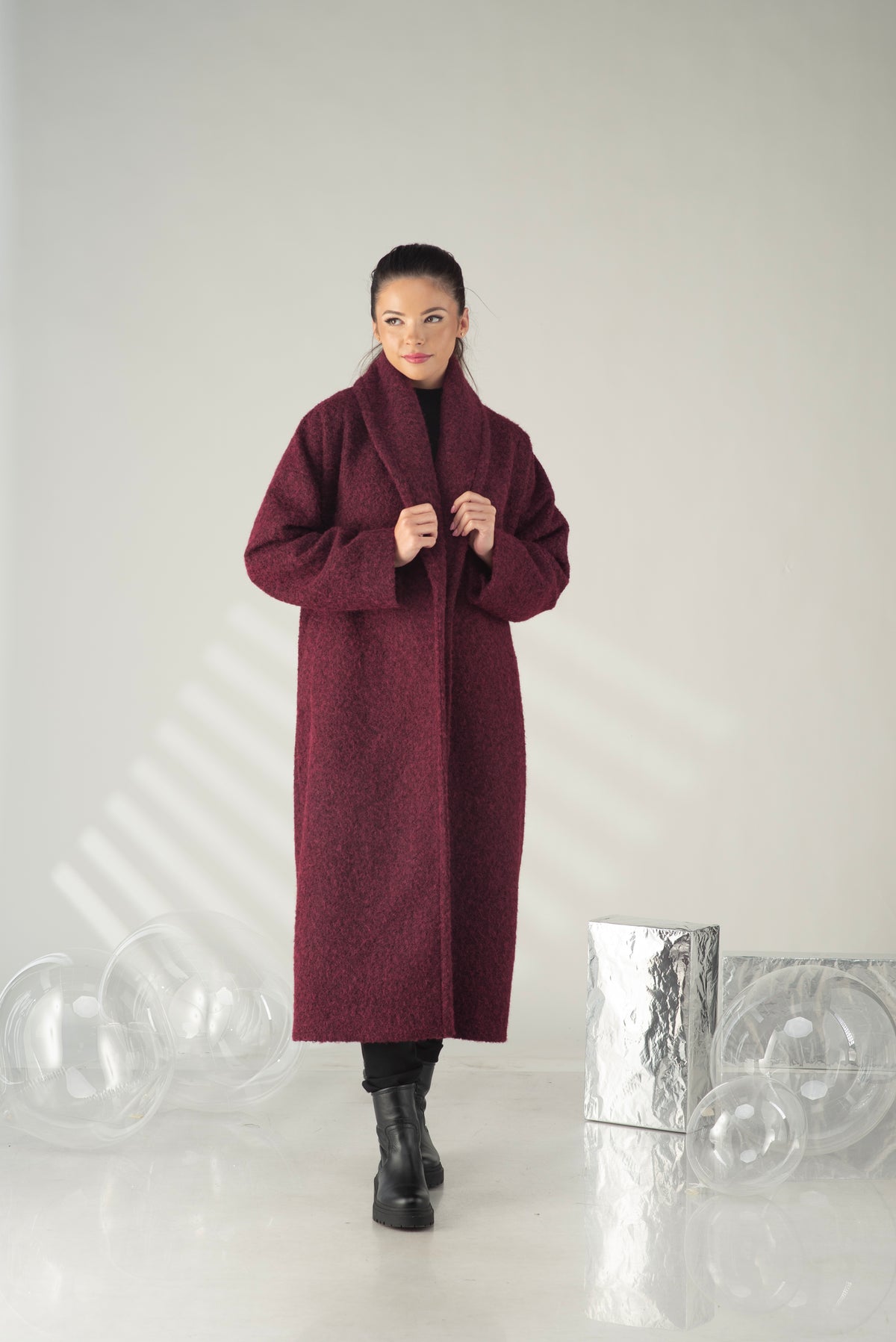 A woman modeling a long, oversized maroon coat with a high shawl collar over a black outfit, standing in a minimalist studio with decorative clear glass spheres and a metallic silver box.