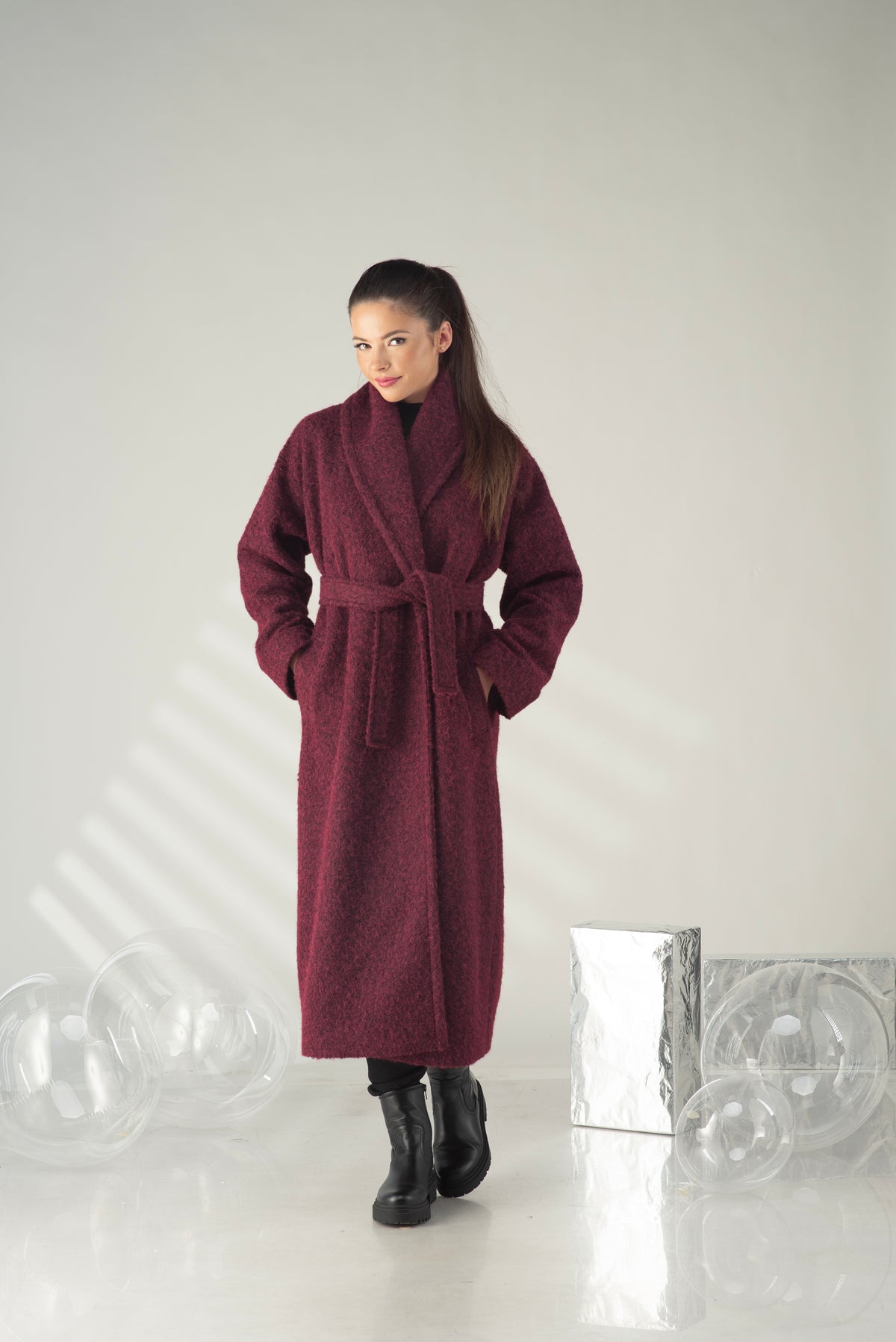 A woman wearing a long, plush maroon bathrobe with a tie belt, standing in a studio setting with a light gray background and decorative clear glass spheres.