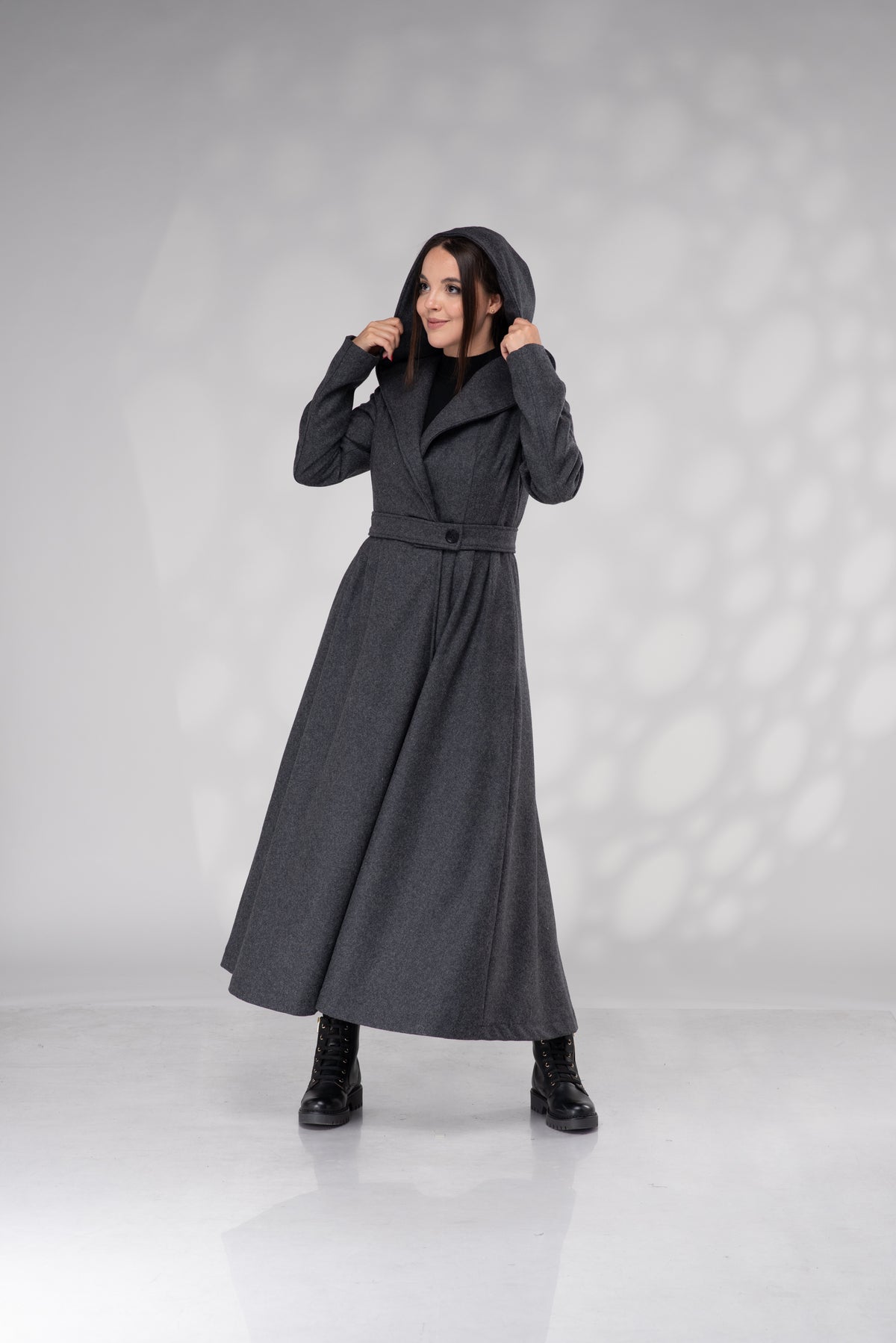 A woman modeling a long, dark gray hooded coat with a belted waist, standing against a light gray studio backdrop with soft shadow patterns.