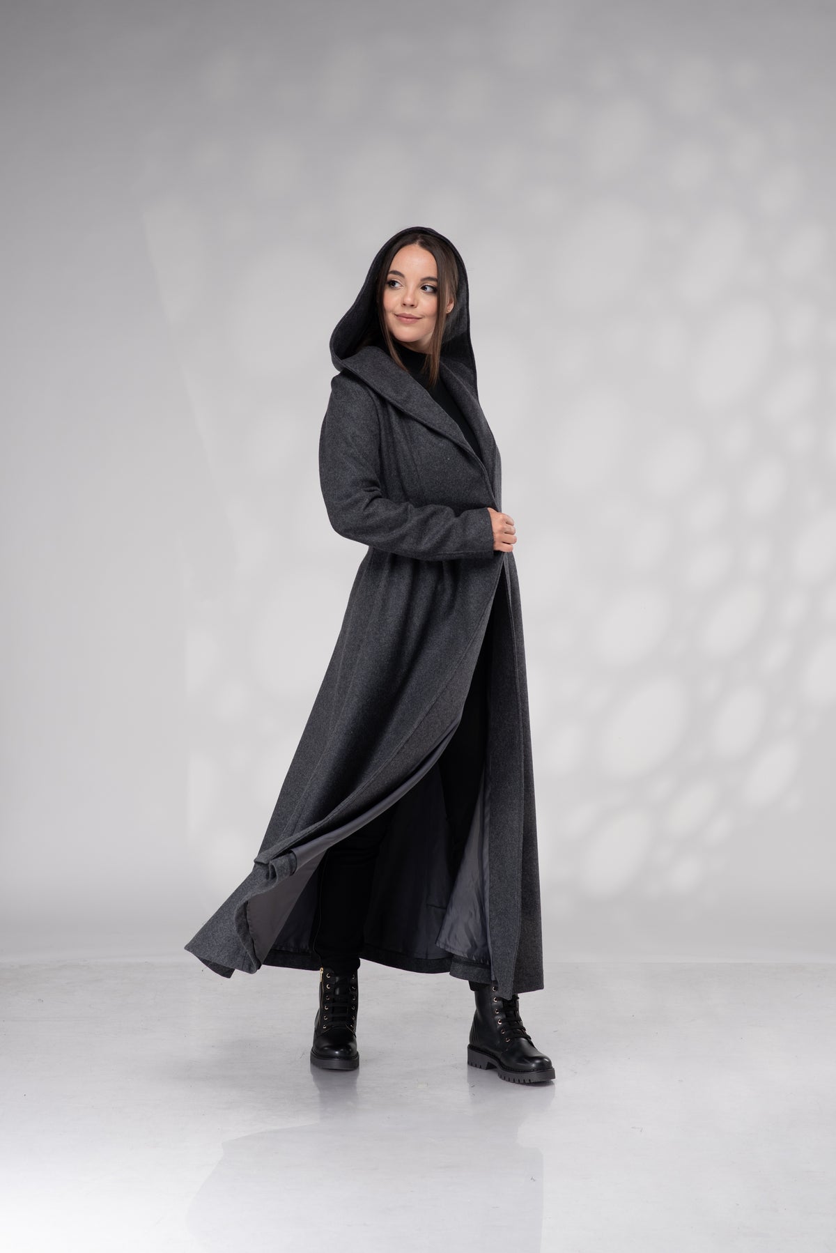 A woman modeling a long, dark charcoal gray hooded coat with a wide collar, standing in a studio with a light gray background and soft shadow patterns.