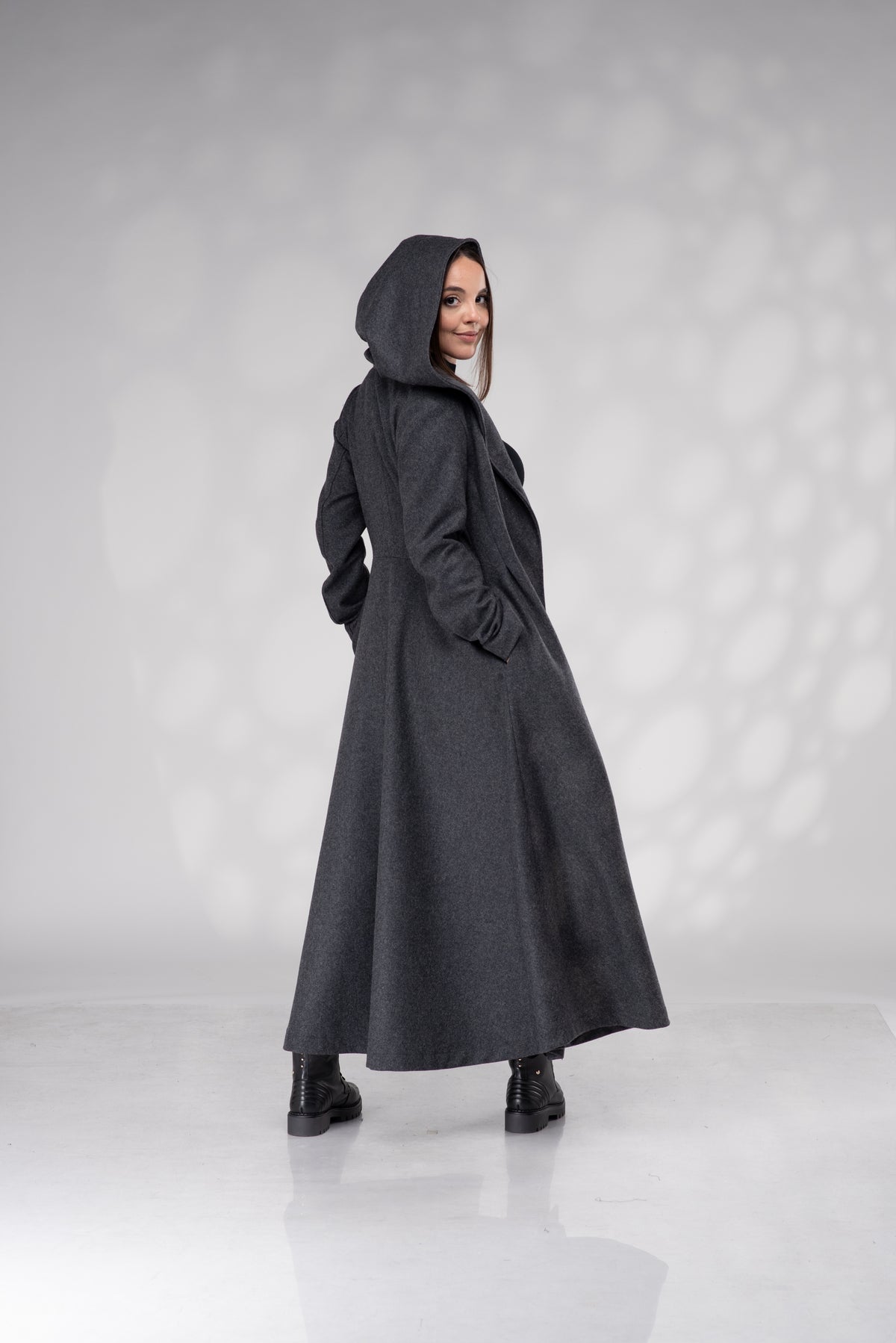 A woman wearing a long, dark gray hooded coat with pockets, standing in a studio with a light gray background and soft shadow patterns.
