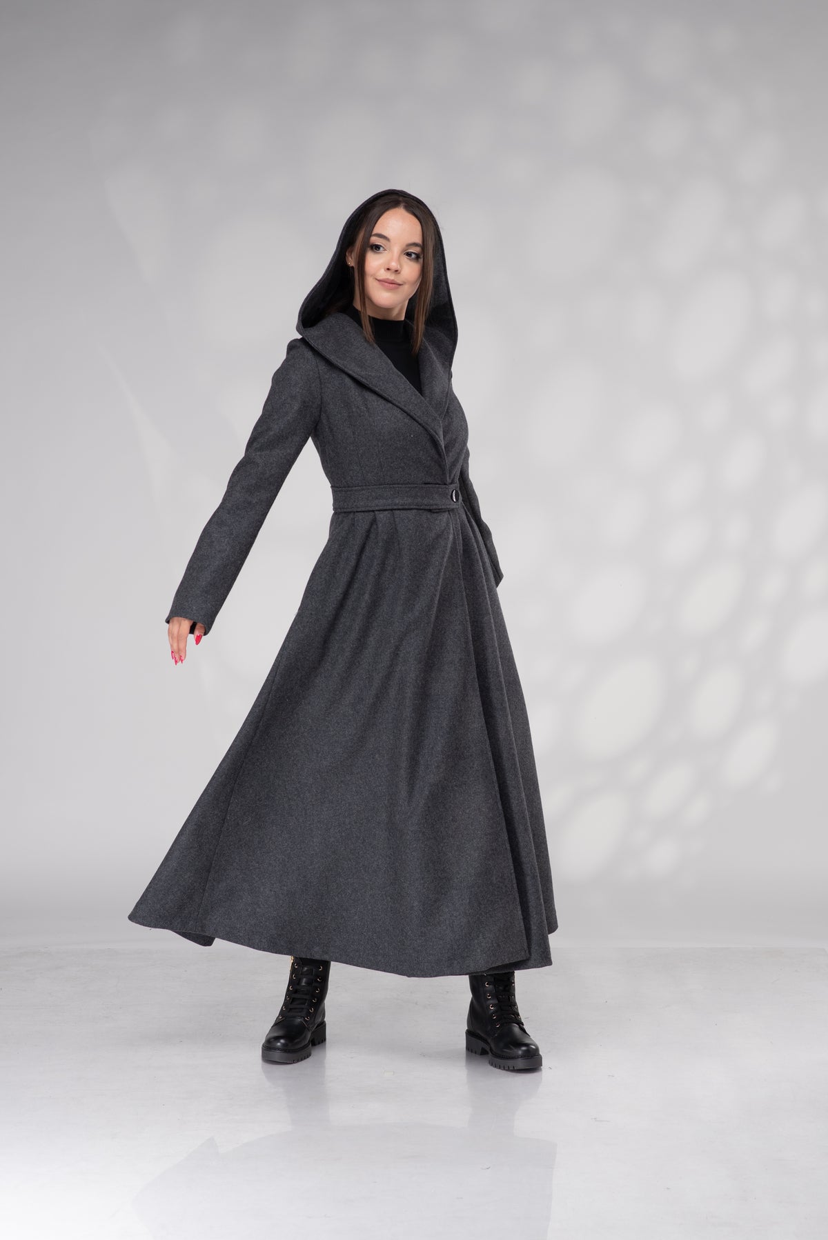 A woman modeling a long, dark gray wrap-style coat with a hood, worn over a black top and black boots, standing against a light gray studio backdrop with soft shadow patterns.