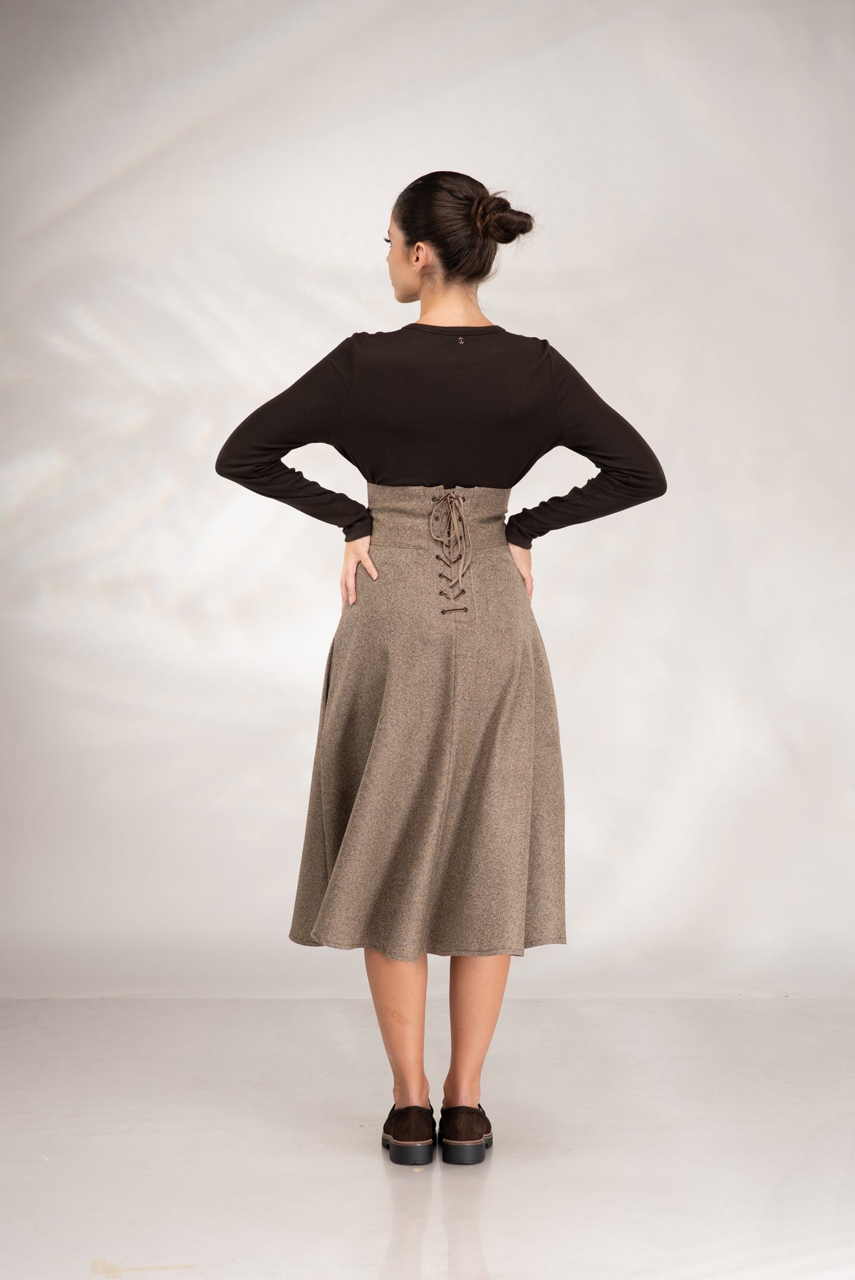 A woman is shown from the back wearing a black long-sleeve top and a high-waisted taupe brown skirt with lace-up detailing at the back waist, standing against a light gray studio backdrop.