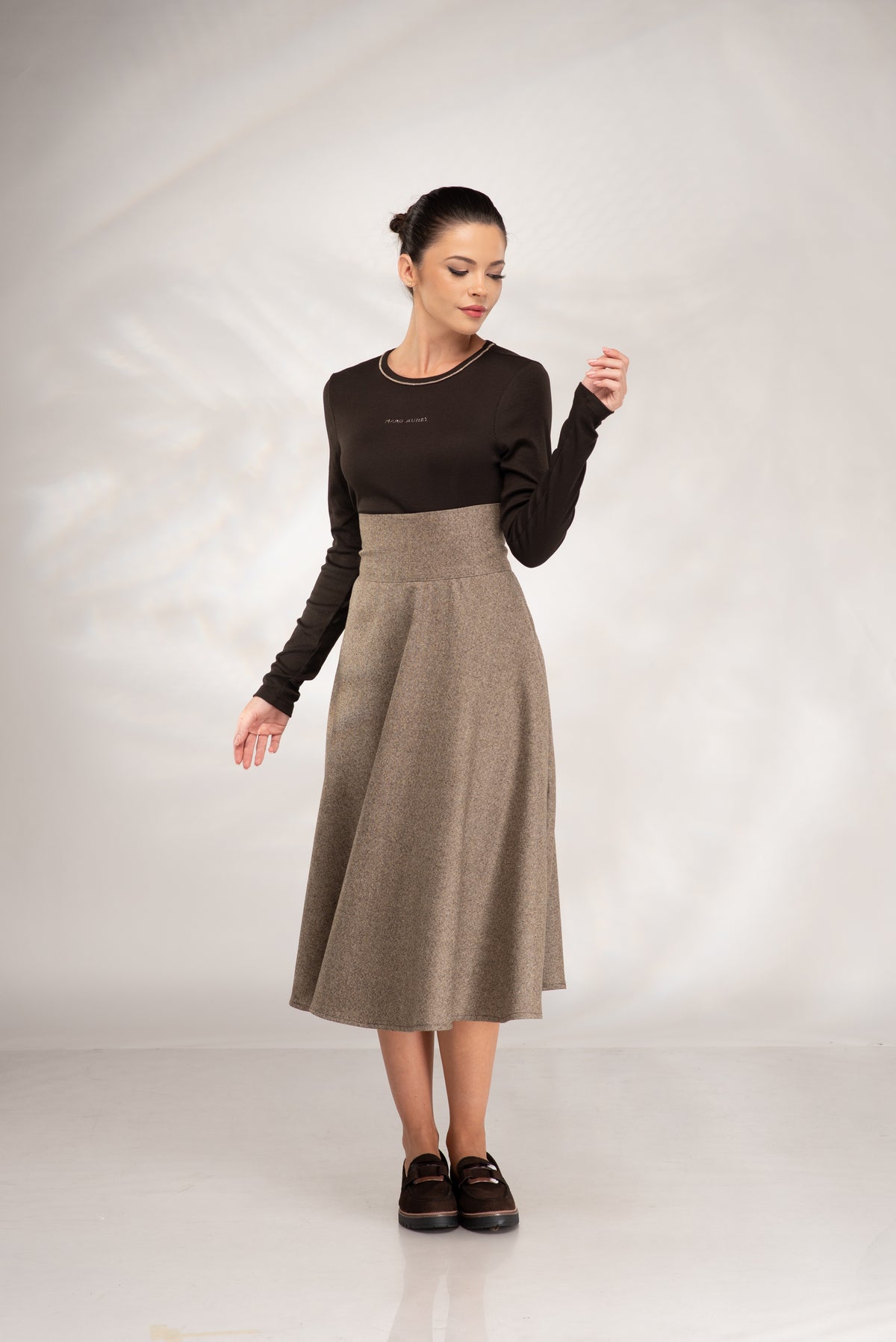 A woman modeling a two-piece outfit consisting of a dark brown long-sleeve knit top and a high-waisted, knee-length taupe tweed skirt, paired with dark brown loafers, against a light gray studio backdrop.